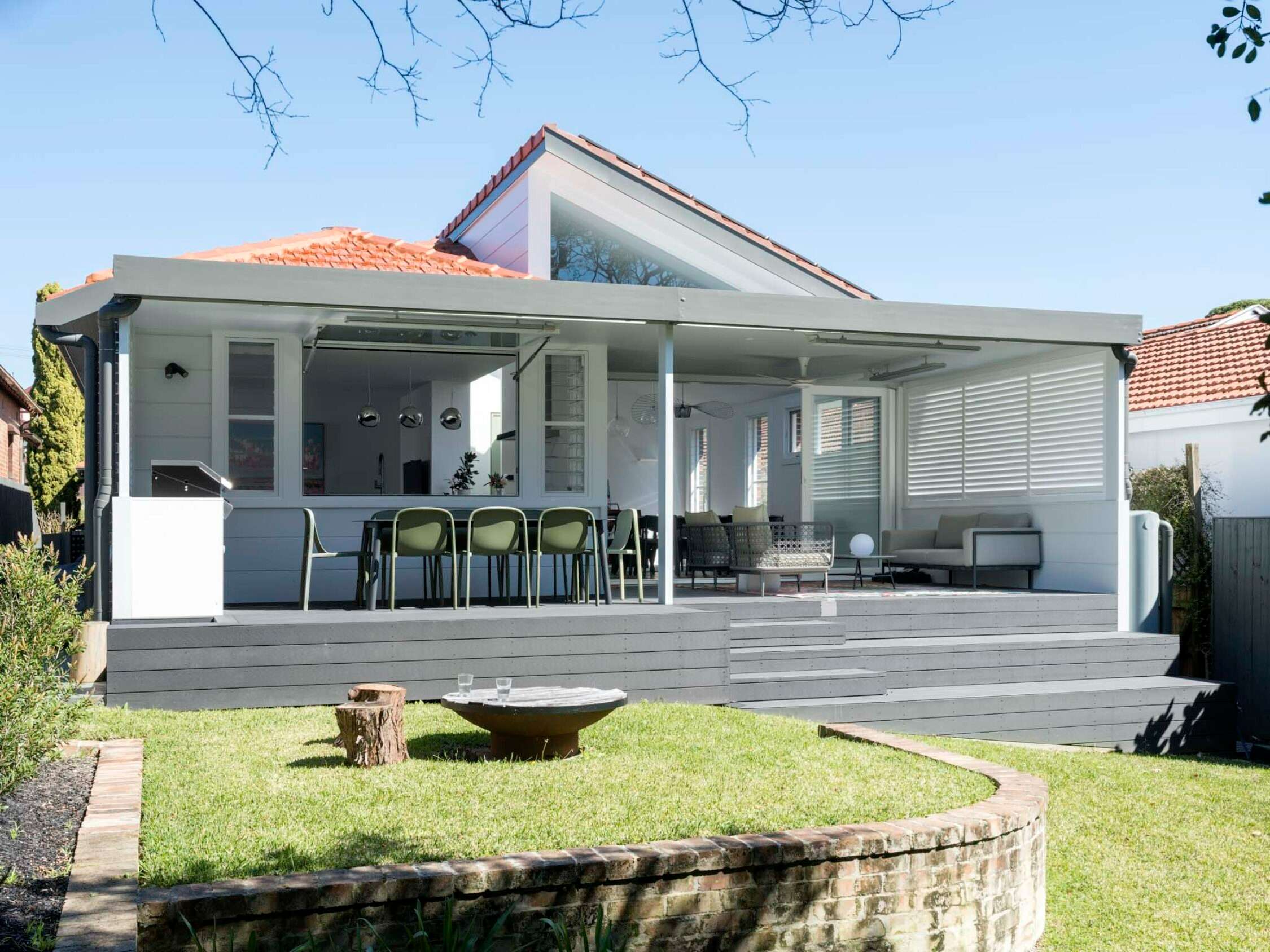 Covered alfresco with composite deck, outdoor dining and lounge areas, folding glass doors, louvred shutters, and a lawned garden with a fire bowl.