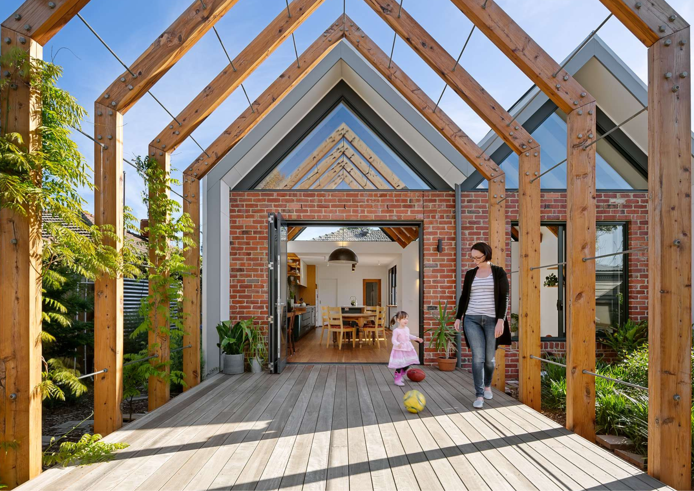 Timber pergola structure with exposed rafters and wire trellis over a composite deck, opening to a brick home interior with a child and adult in the outdoor space.