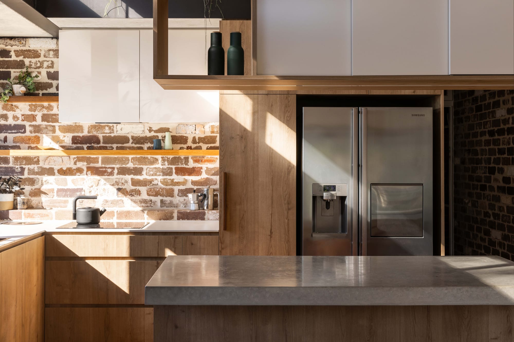 Kitchen detail in Manly by Matt Day Architect showing timber cabinetry, exposed recycled brick splashback, stone benchtop and natural light cast across the surfaces.