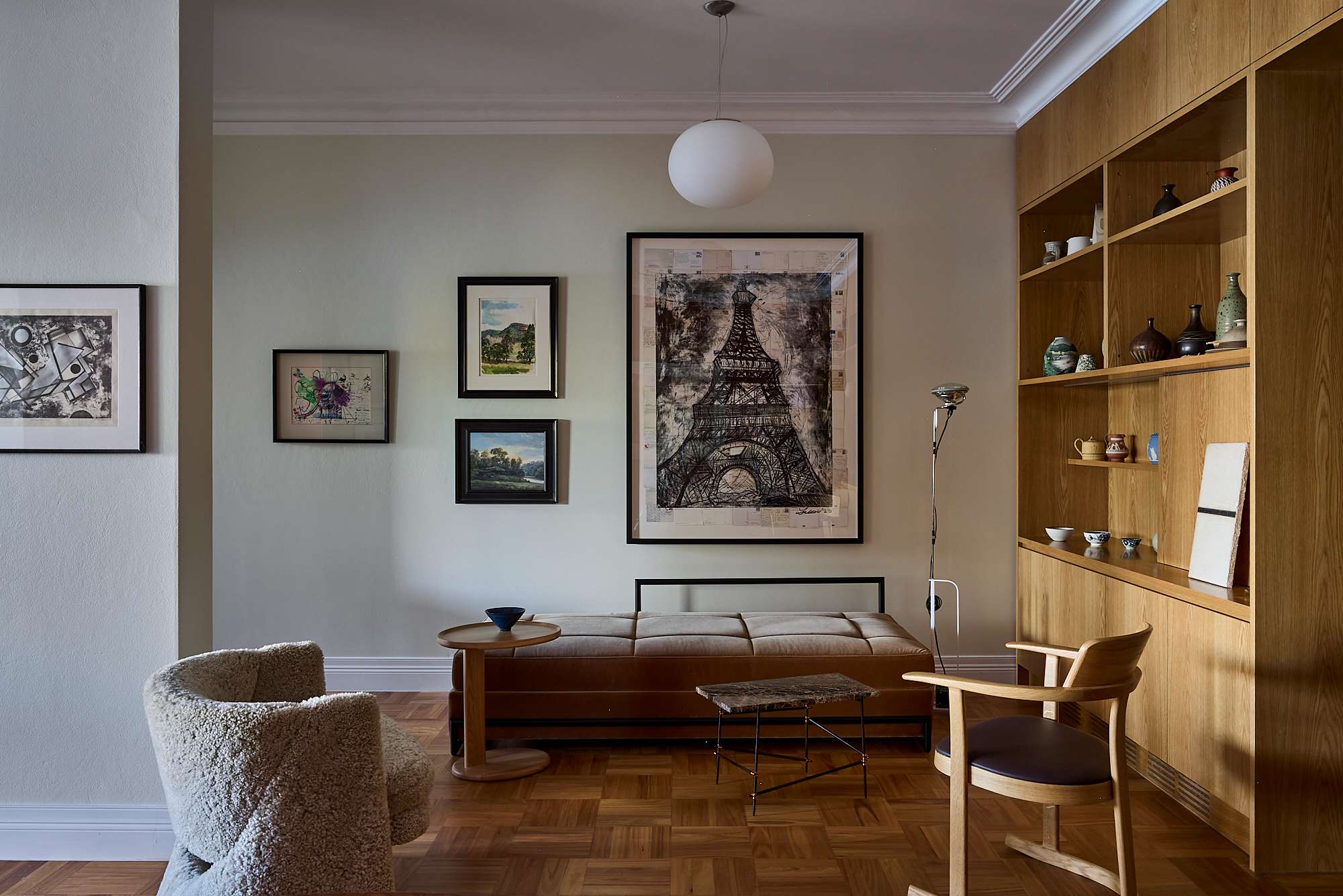 A living room interior featuring a mid-century daybed in tan leather beneath a large framed Eiffel Tower artwork, flanked by a gallery wall of mixed-media prints, a shearling armchair, an oak shelving unit displaying ceramic vessels, and parquet timber flooring.