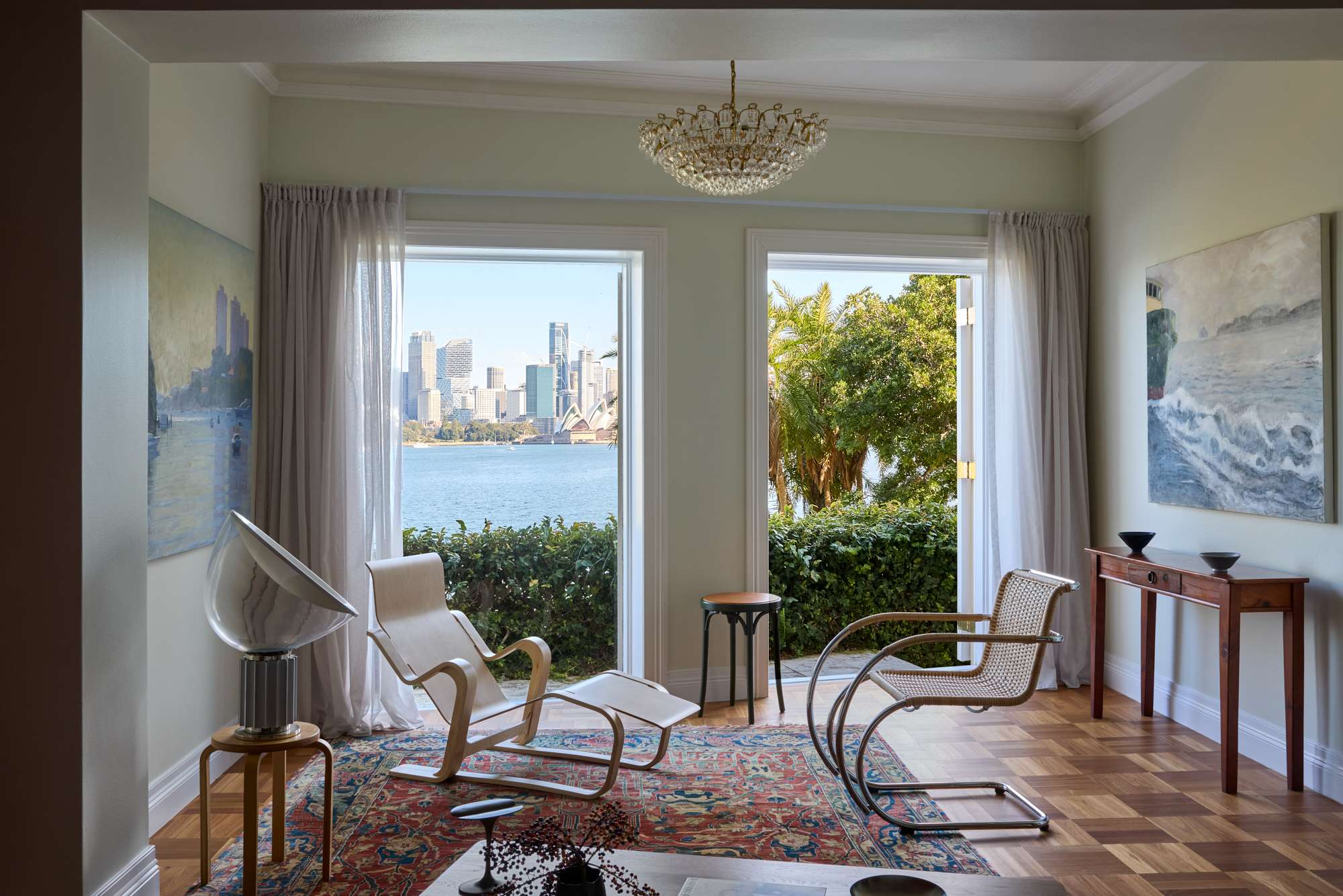 A light-filled sitting room with direct harbour views of the Sydney CBD and Opera House through full-height windows, furnished with an Aalto lounge chair and ottoman, a Mies van der Rohe cantilever chair, a Persian rug, and a vintage crystal chandelier overhead.
