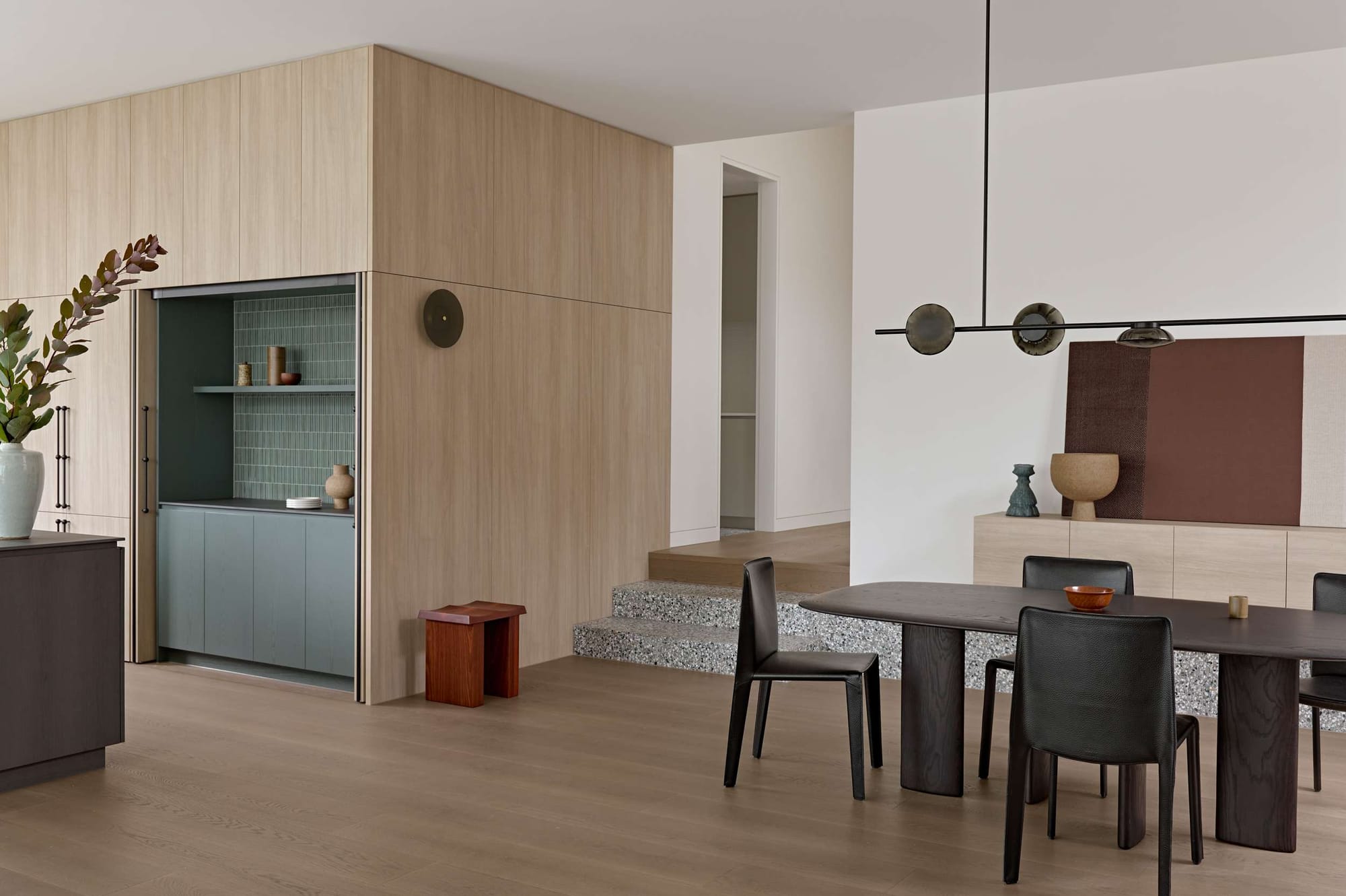 Open-plan dining and living area in Orrong House by Studio Cobe, with concealed green-tiled joinery, terrazzo stepped platform, oak cabinetry and a dark timber dining setting.