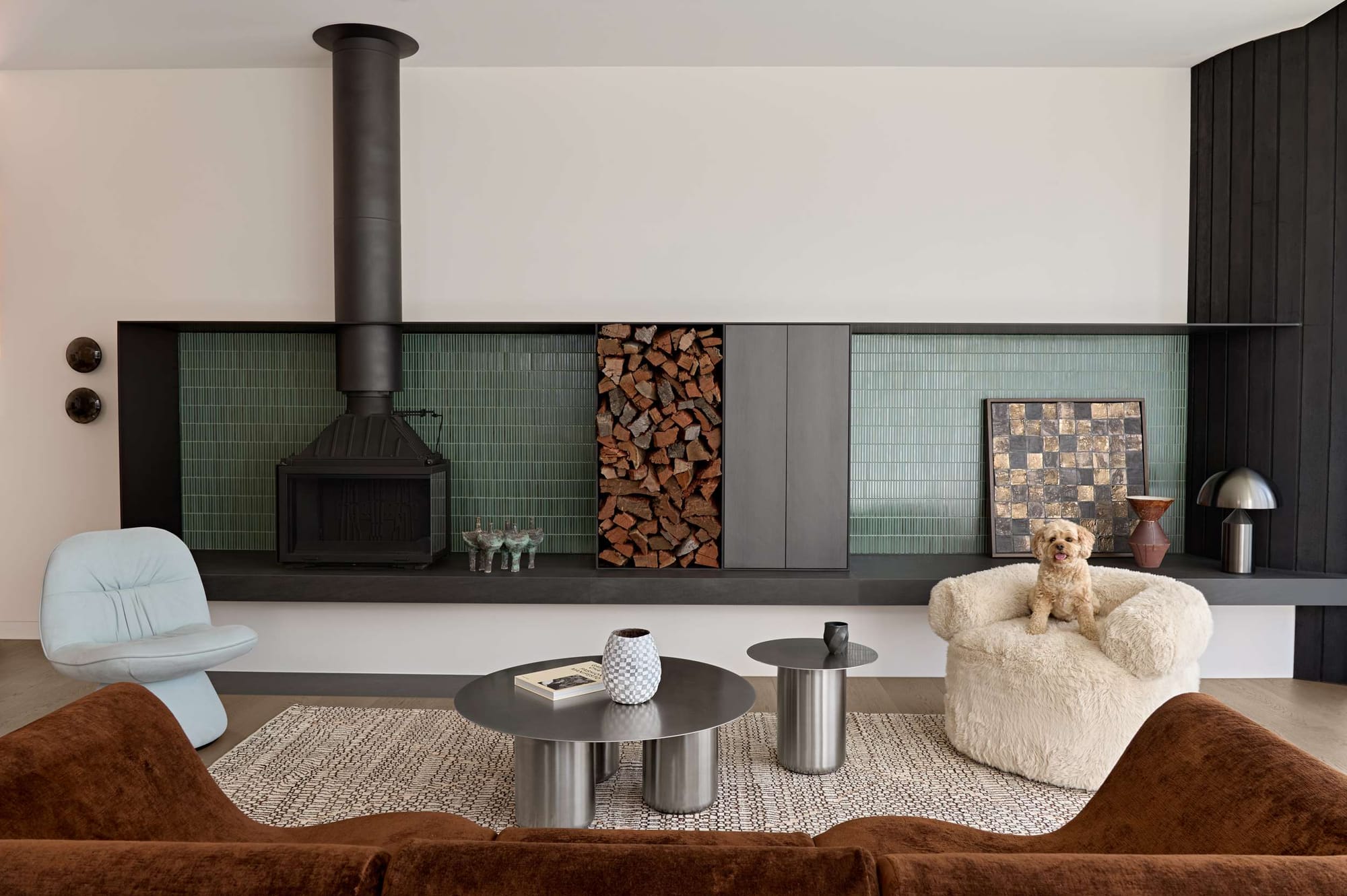 Living room of Orrong House by Studio Cobe featuring a cast-iron freestanding fireplace, green tiled joinery wall, curved furniture and a small dog seated in a boucle armchair.