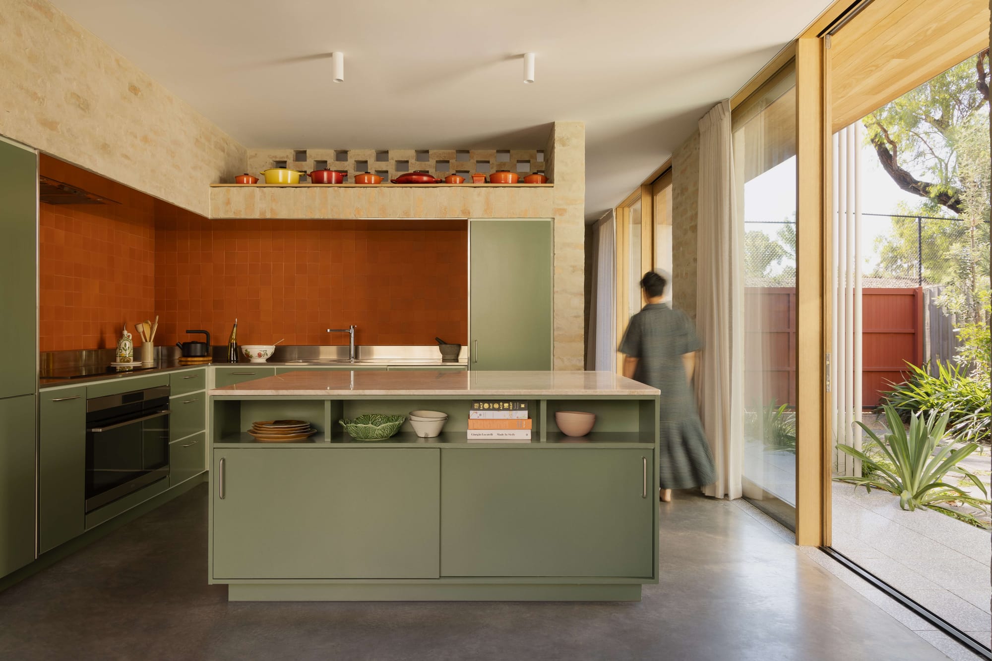 Sage green kitchen with terracotta tile splashback, stone walls and sliding doors opening to courtyard garden, Oval House by Topology Studio