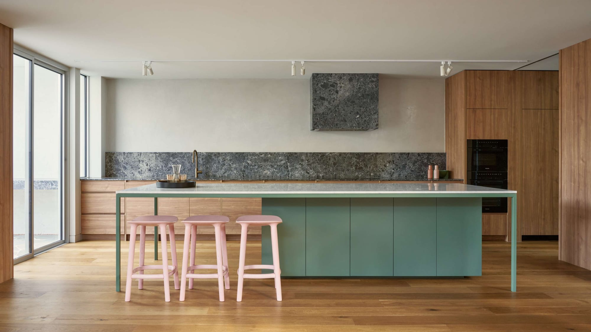 Kitchen of Peavey Residence II by FYC Architects with a sage green island, pink timber stools, stone splashback and warm timber joinery throughout