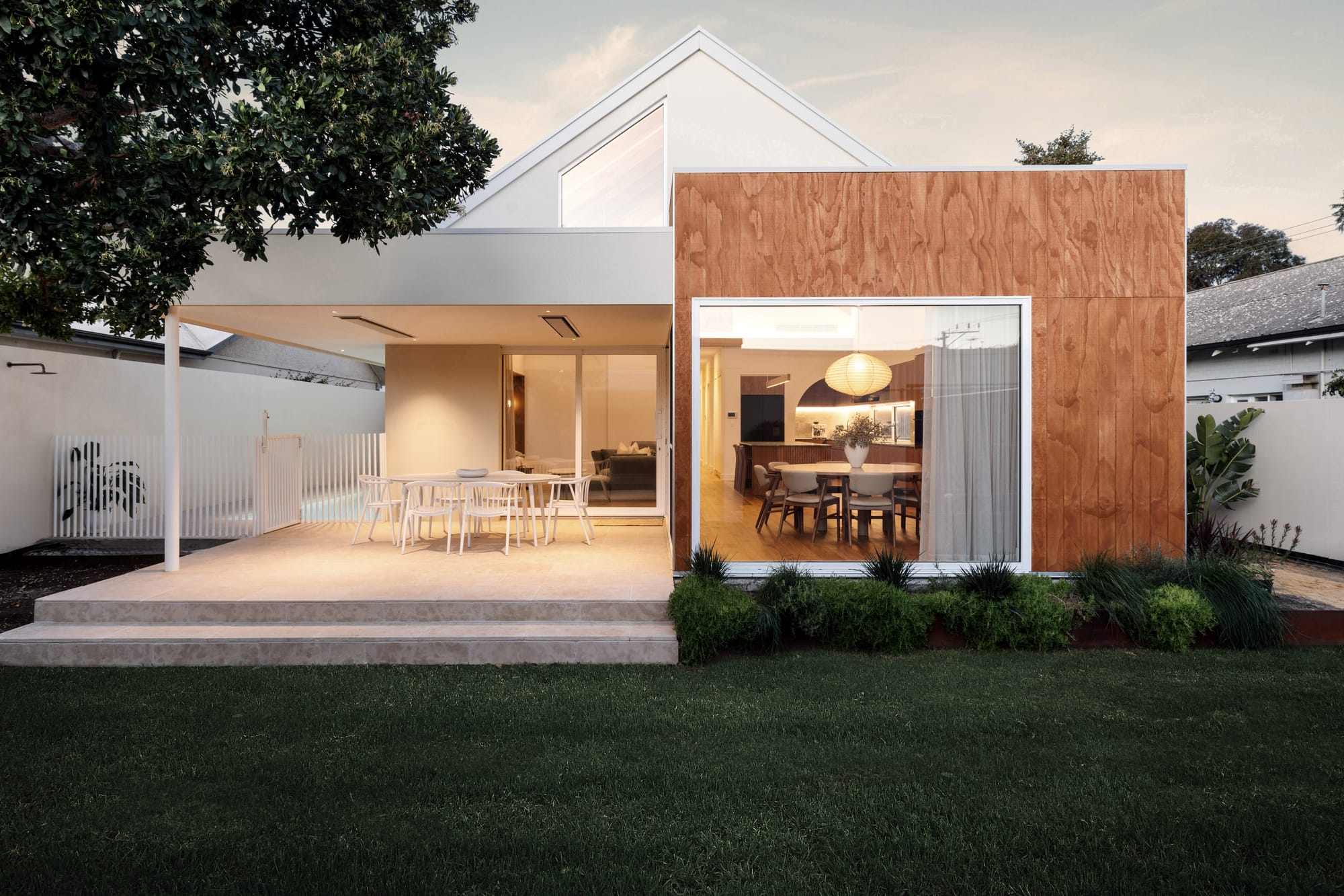 Rear facade of Playhouse by Trennel Architecture at dusk, showing a timber-clad extension with large glazed opening, covered outdoor entertaining area and garden lawn.