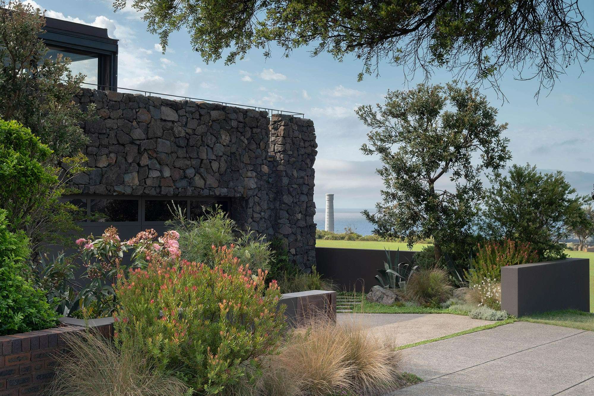 Dense native planting including grevilleas and ornamental grasses frames a path alongside a rugged stone clad home with ocean views beyond, Stone House by Secret Gardens