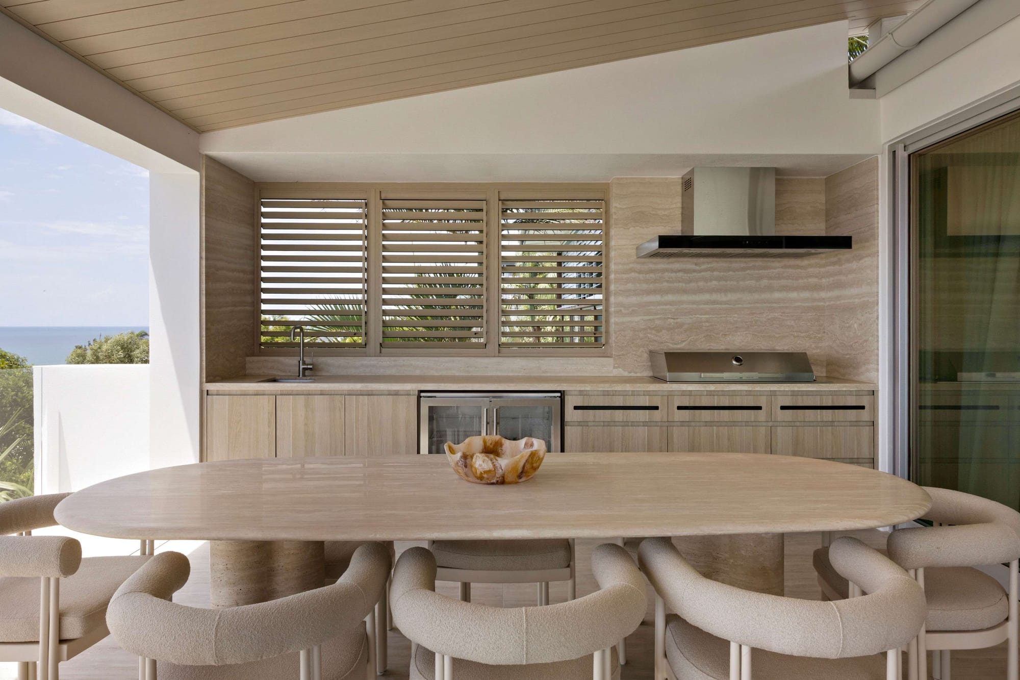 Premium covered outdoor kitchen with stone benchtops, integrated barbecue, underbench refrigeration, louvred shutters, and ocean views beyond.