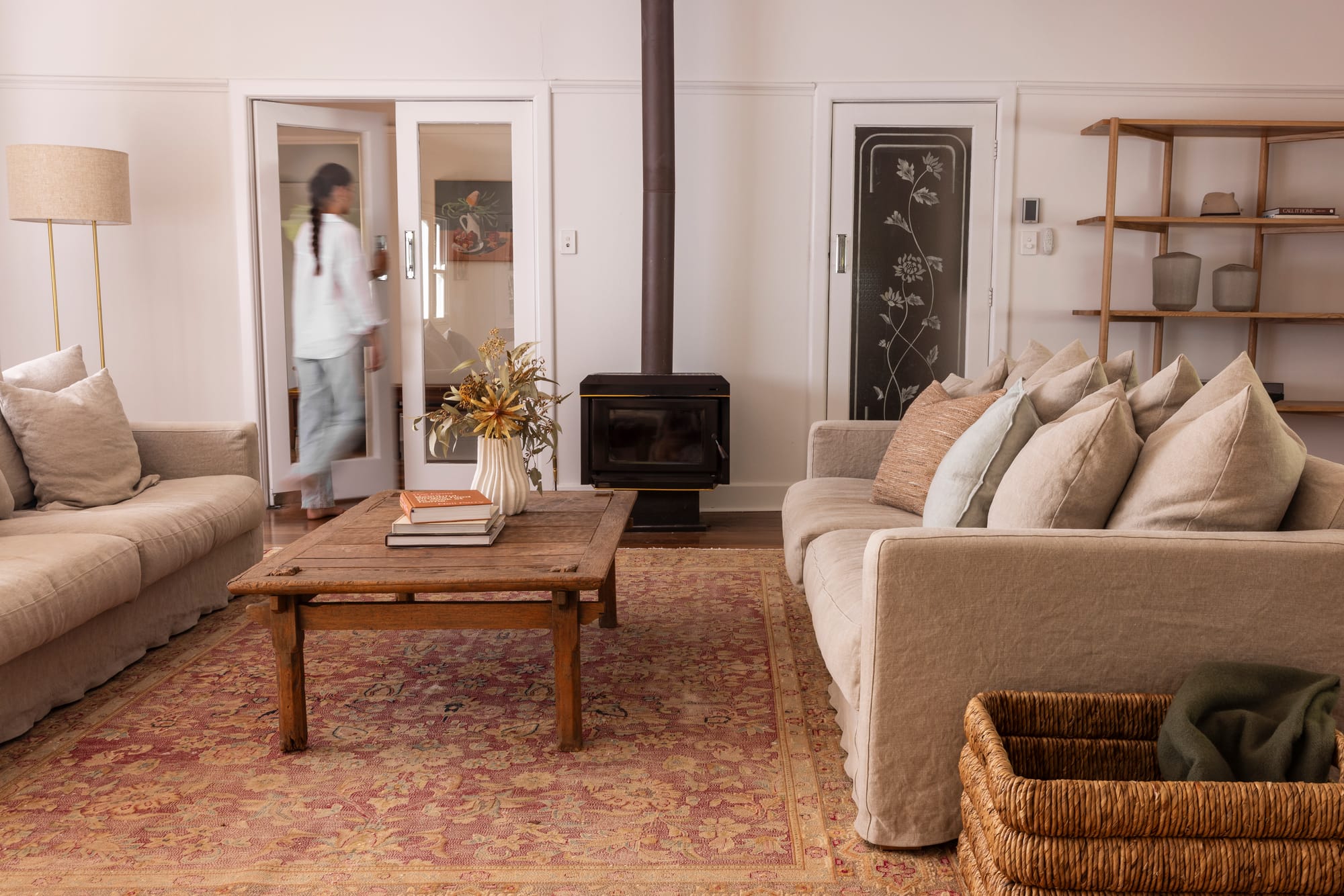 Relaxed heritage cottage living room with two linen sofas, a vintage timber coffee table, a patterned Persian rug, a freestanding wood heater, etched glass panel doors, and open timber shelving styled with ceramics, with a motion-blurred figure passing through a doorway.