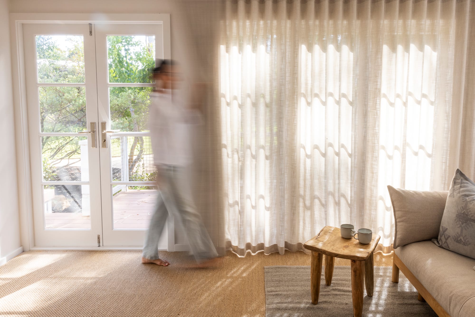 Light-filled living space with sheer linen curtains diffusing afternoon sun, a motion-blurred figure moving toward open French doors, a rustic timber side table with two mugs, and a linen sofa with textured cushions.