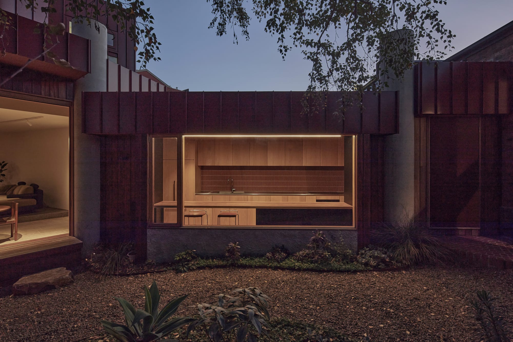 A dusk view into a warmly lit outdoor kitchen framed by a dark corrugated steel and concrete facade, with timber cabinetry, strip lighting, and a gravel courtyard garden in the foreground.
