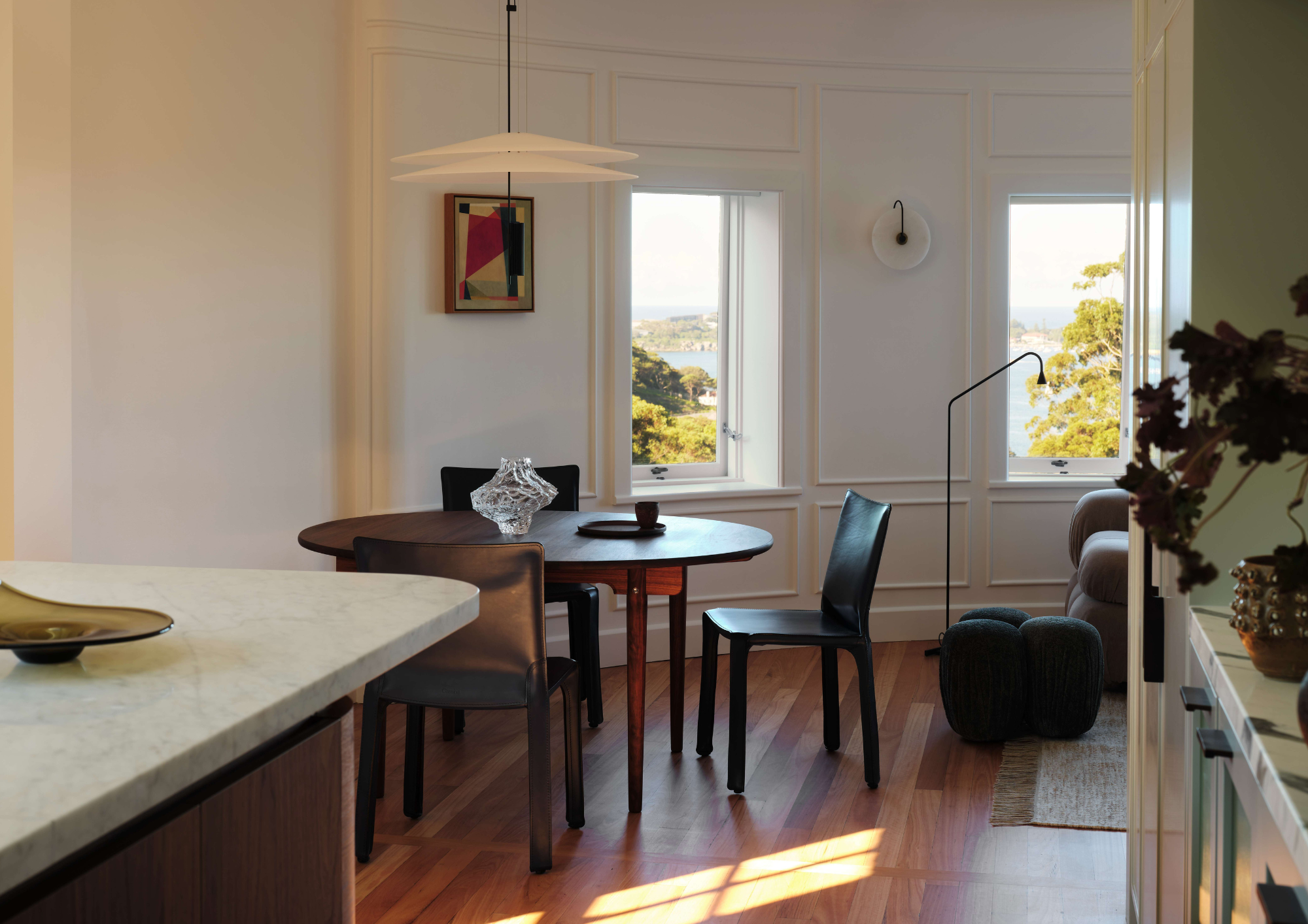 A warm dining area with a round dark timber table, black leather chairs, and a flat pendant light, set against white panelled walls with harbour views through double windows.