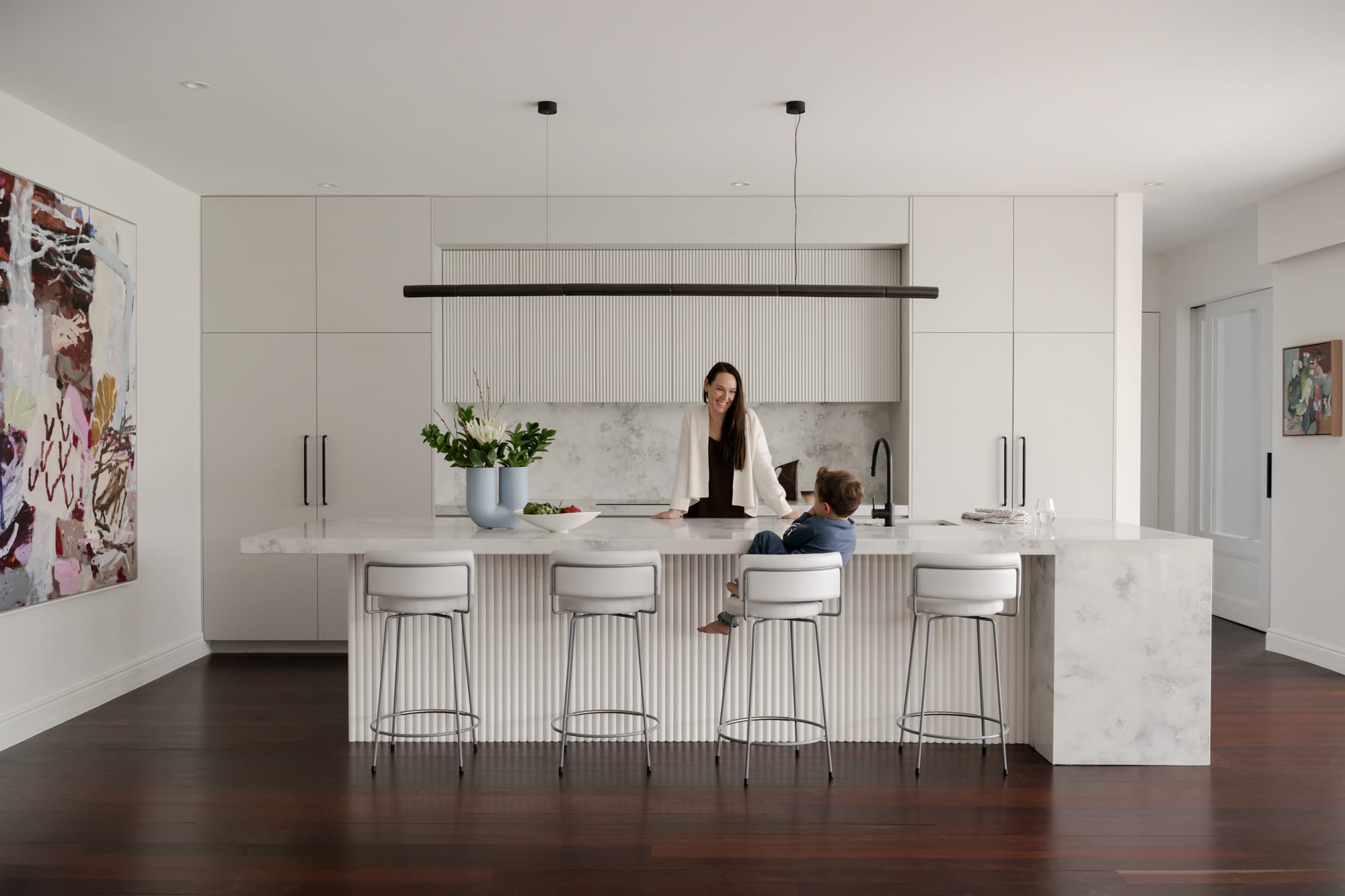 A mother and child at a fluted marble kitchen island in a Keen Architecture home, with dark timber flooring and a large abstract artwork on the wall.