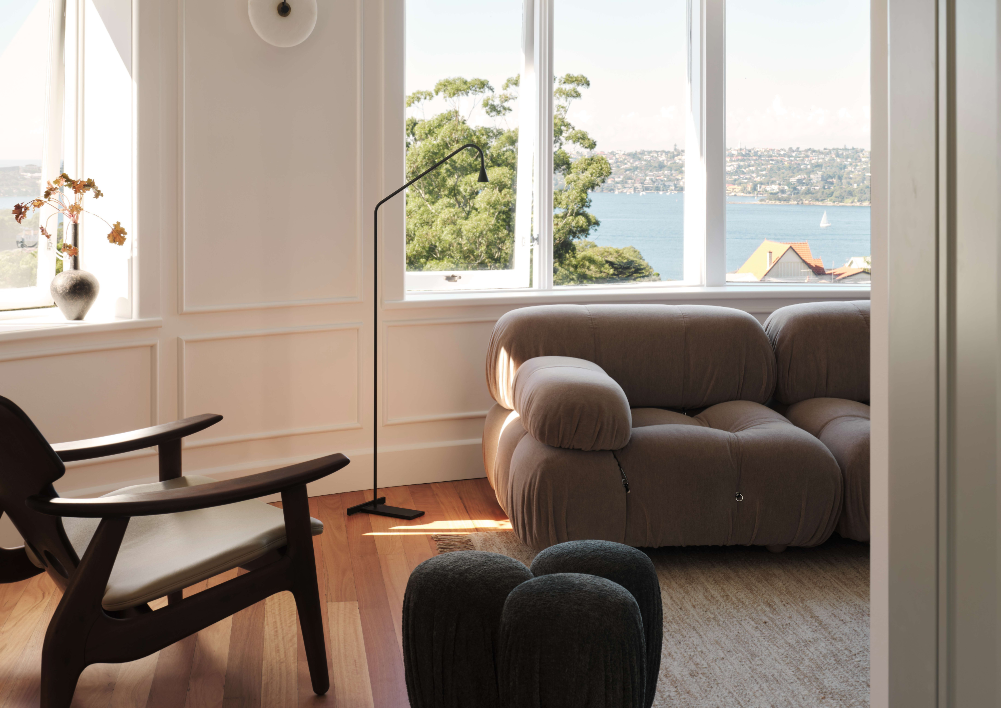 A curved living room corner with a sculptural taupe bouclé sofa, dark timber armchair, black floor lamp, and textured ottomans, framed by white panelled walls and wide windows overlooking Sydney Harbour.