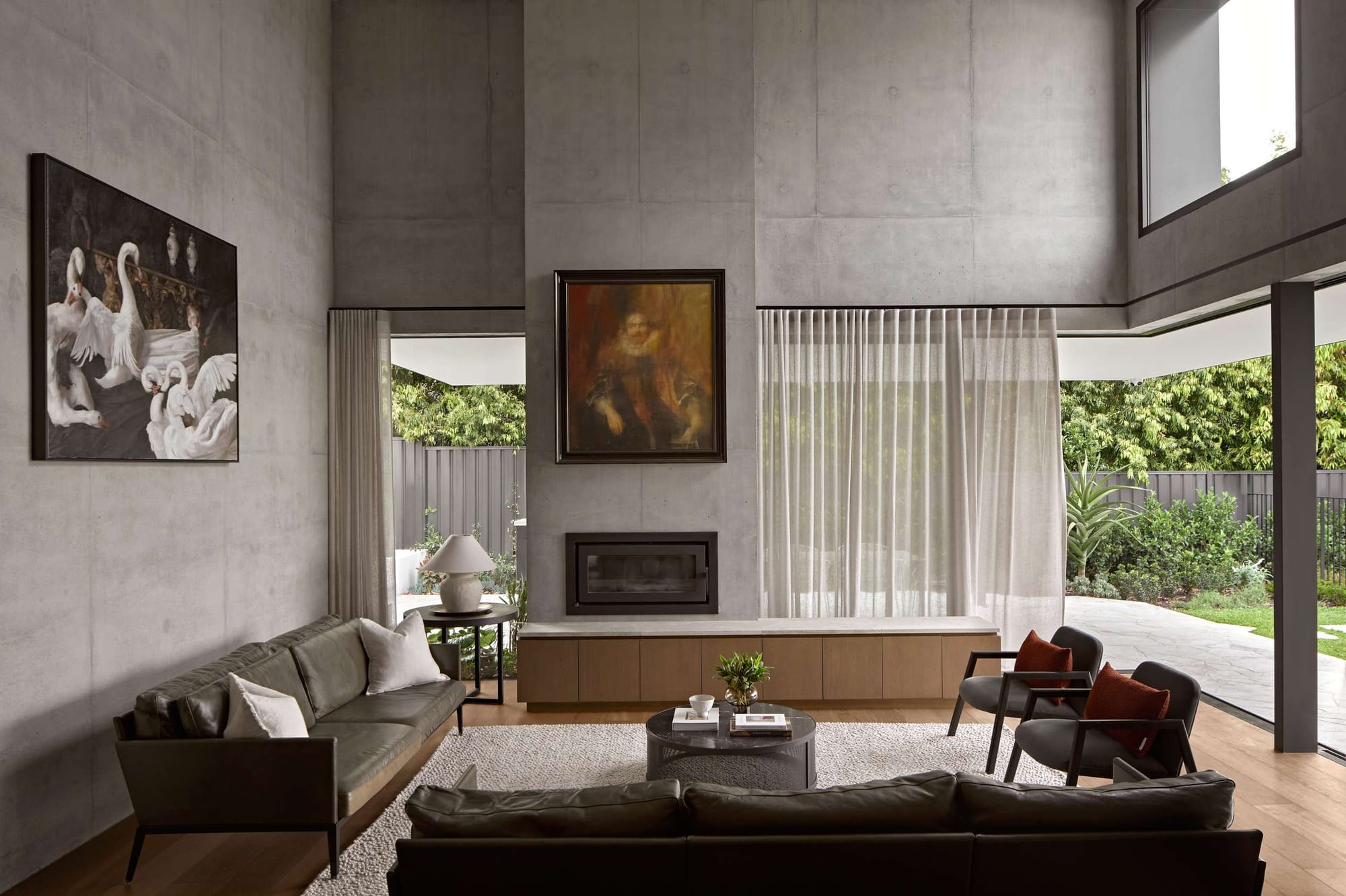 Living room with exposed concrete walls, a classical painting of swans and a portrait hung above a built in fireplace, dark leather sofas and armchairs with rust cushions arranged on a pale rug. Interior design by Cast Studio. Photography by Jack Lovel.