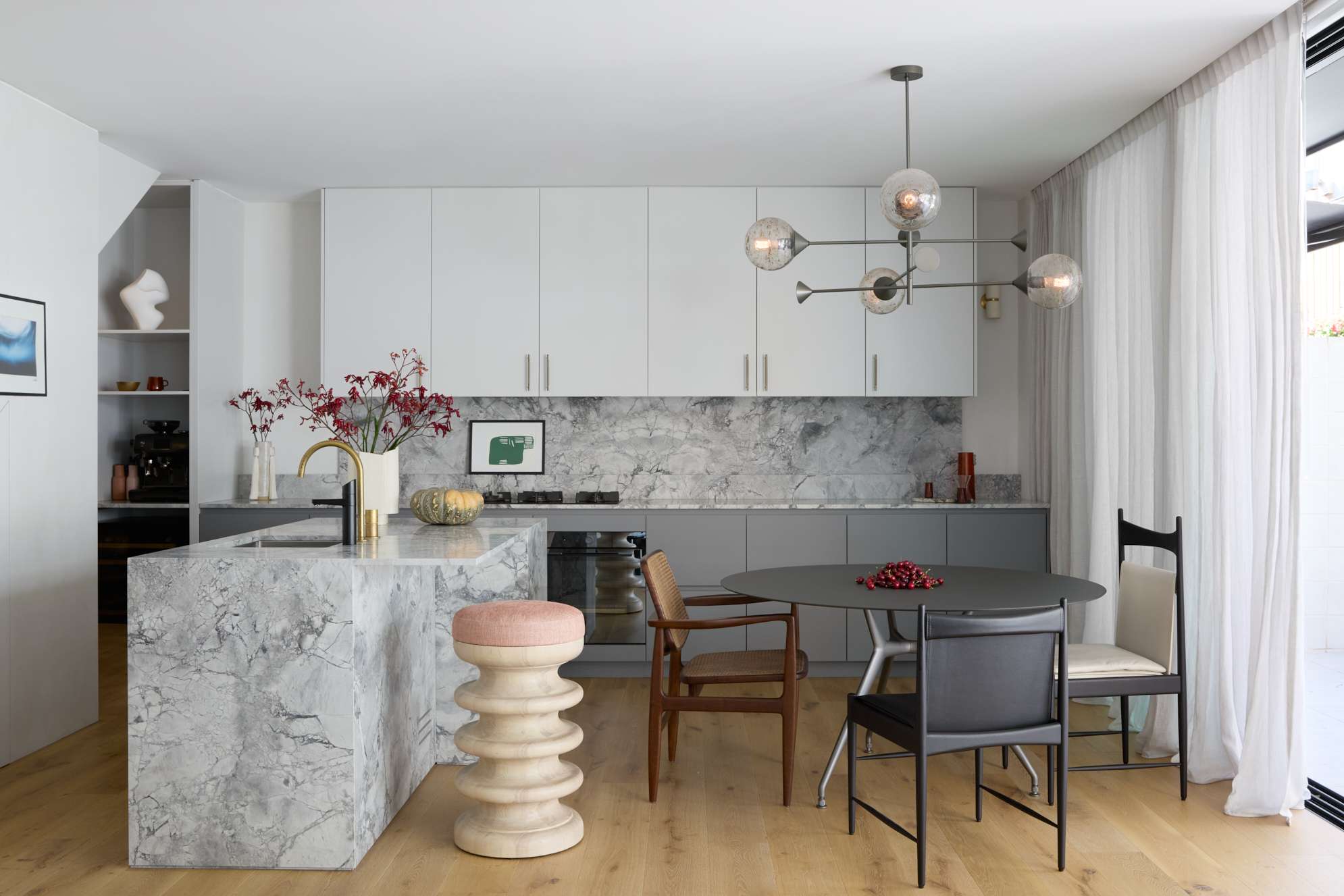 An open-plan kitchen and dining area with a marble waterfall island, white upper cabinetry, grey lower cabinets, a veined stone splashback, sculptural stool, branching glass pendant light, and a round dark dining table with mixed vintage chairs on oak flooring.