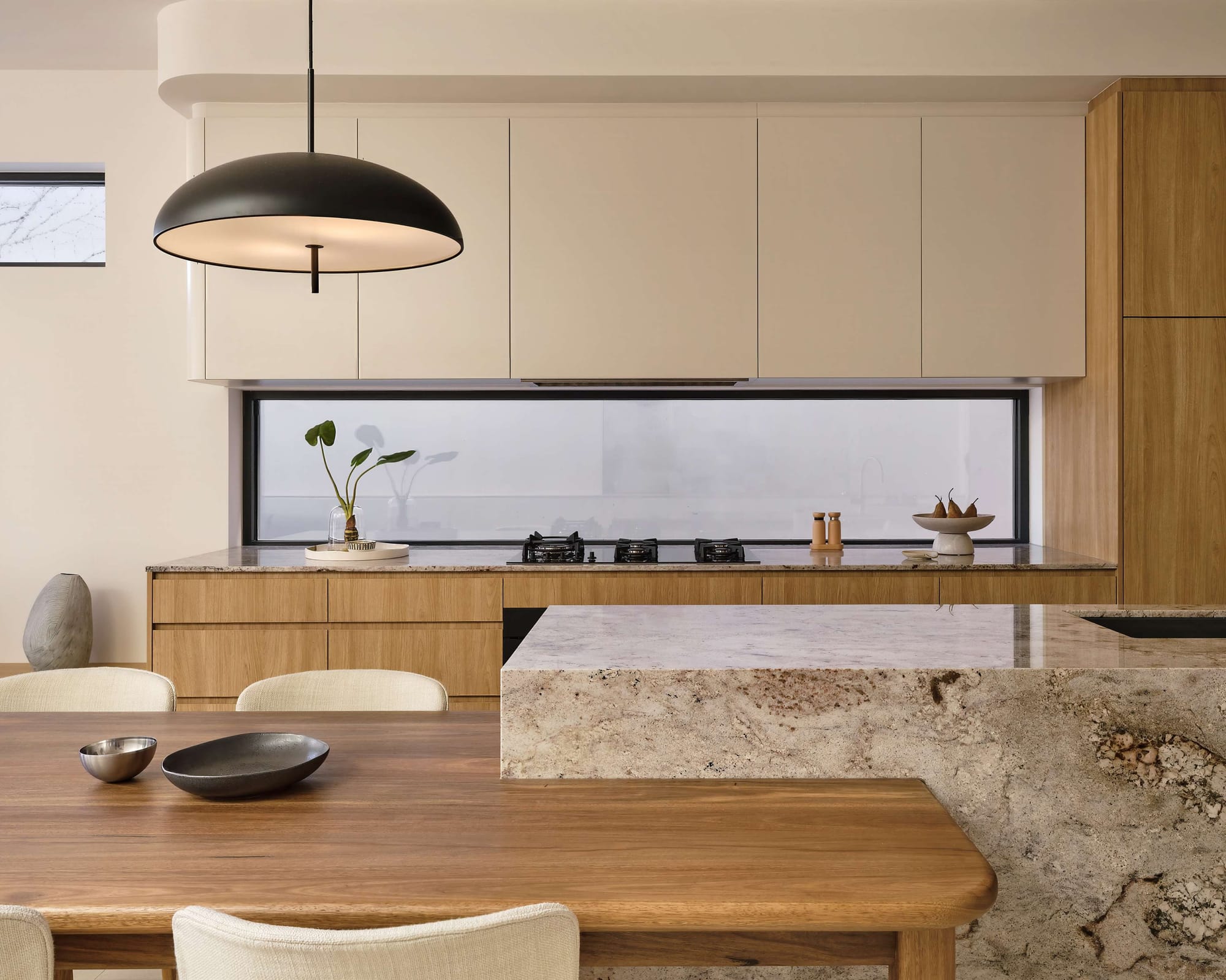 Kitchen with warm timber cabinetry, a textured stone island benchtop, black dome pendant light, and a long window splashback.