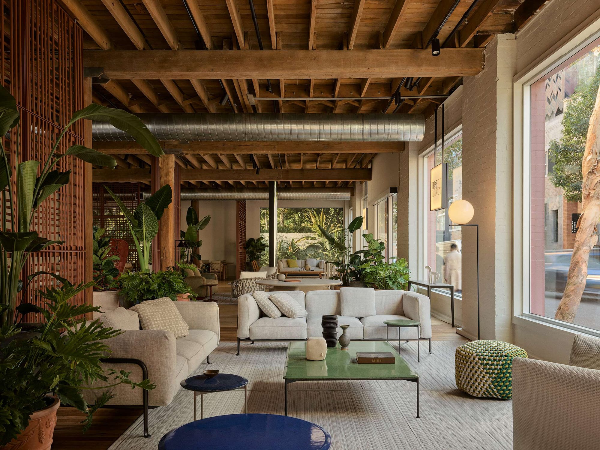 Showroom interior with exposed timber beam ceiling, terracotta screening, and layered neutral toned sofas and coffee tables among lush indoor plants.