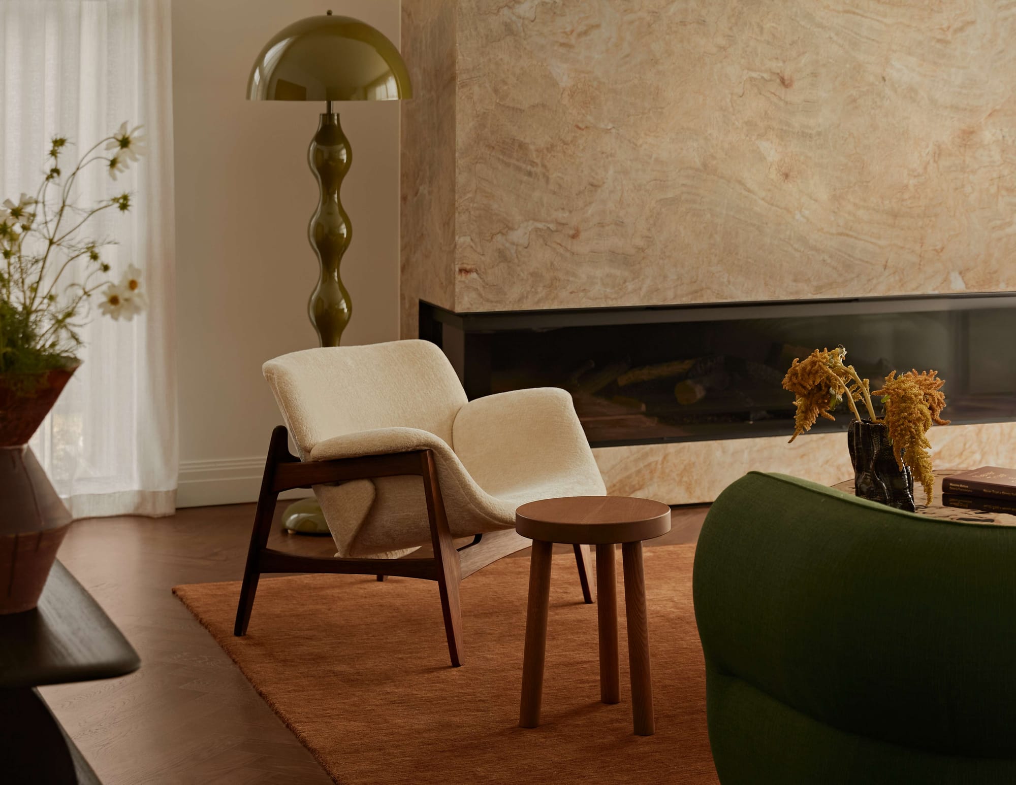 Living room corner with a cream boucle armchair, timber side stool, and a travertine clad fireplace against a rust toned rug.