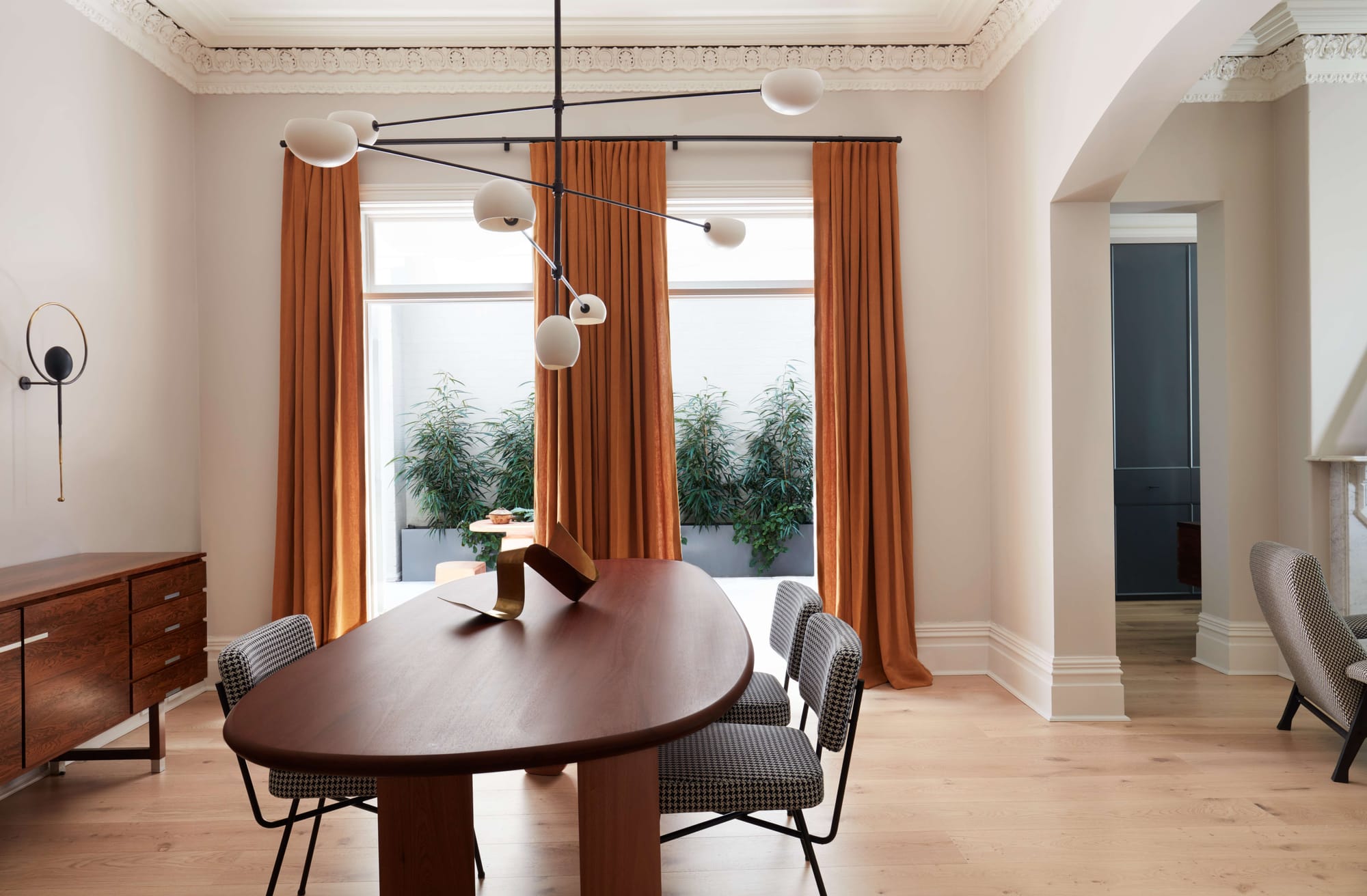 Dining room with ornate ceiling cornicing, burnt orange curtains, a sculptural multi arm pendant light, and a curved timber table with houndstooth chairs.