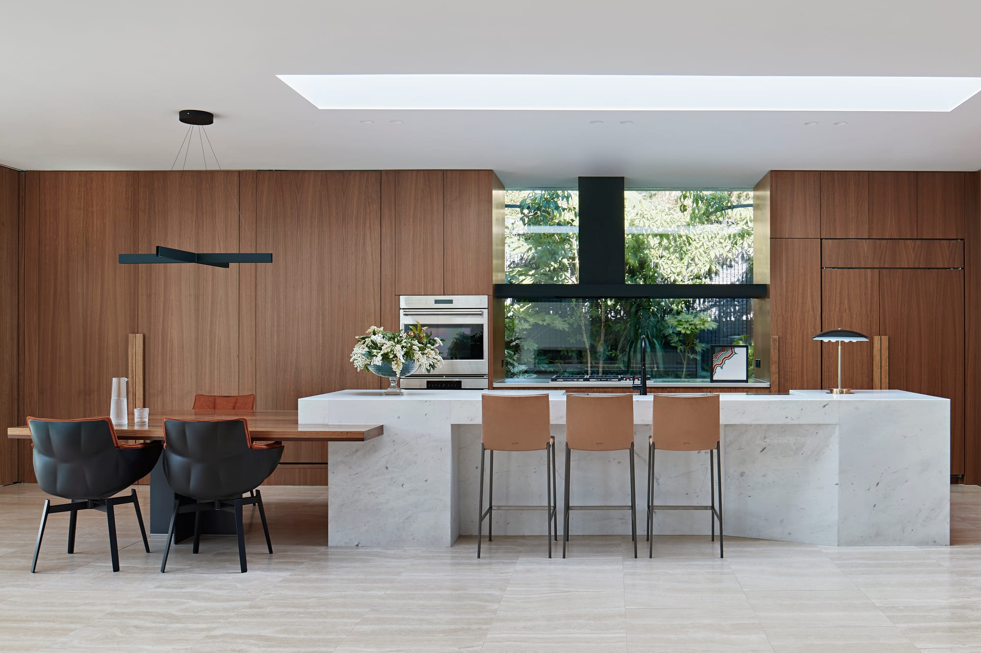 Open plan kitchen and dining space with dark timber cabinetry, a white marble island bench, tan leather bar stools and a timber dining table framed by black leather chairs, with greenery visible through a large window.