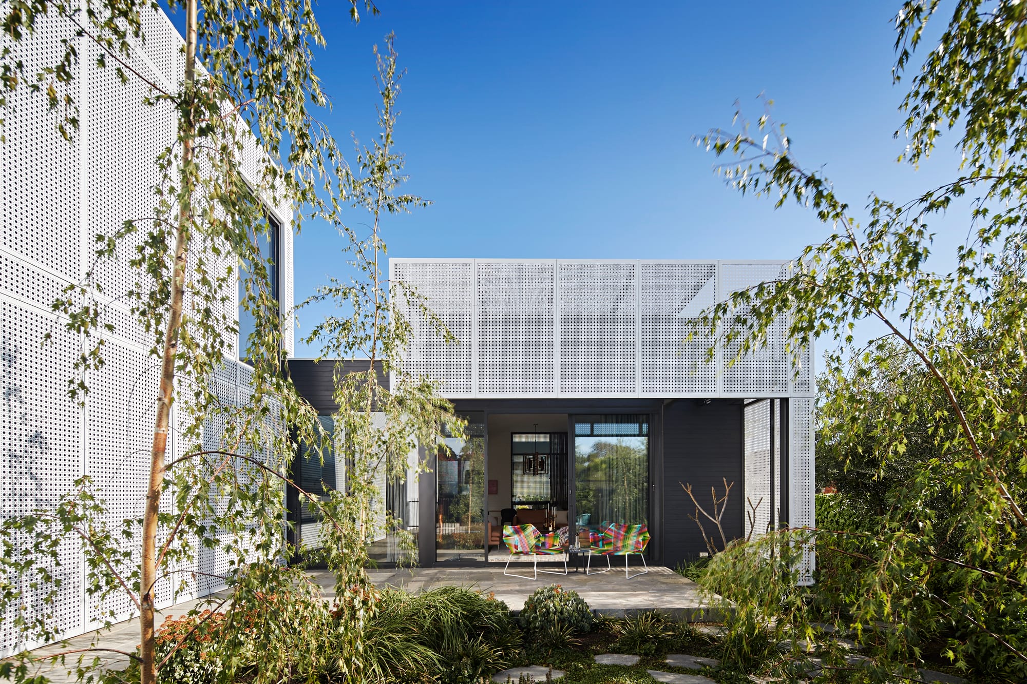 Exterior view of a home with white perforated metal screening and black timber cladding, seen through birch trees, with a paved courtyard and colourful outdoor chairs at the entrance.
