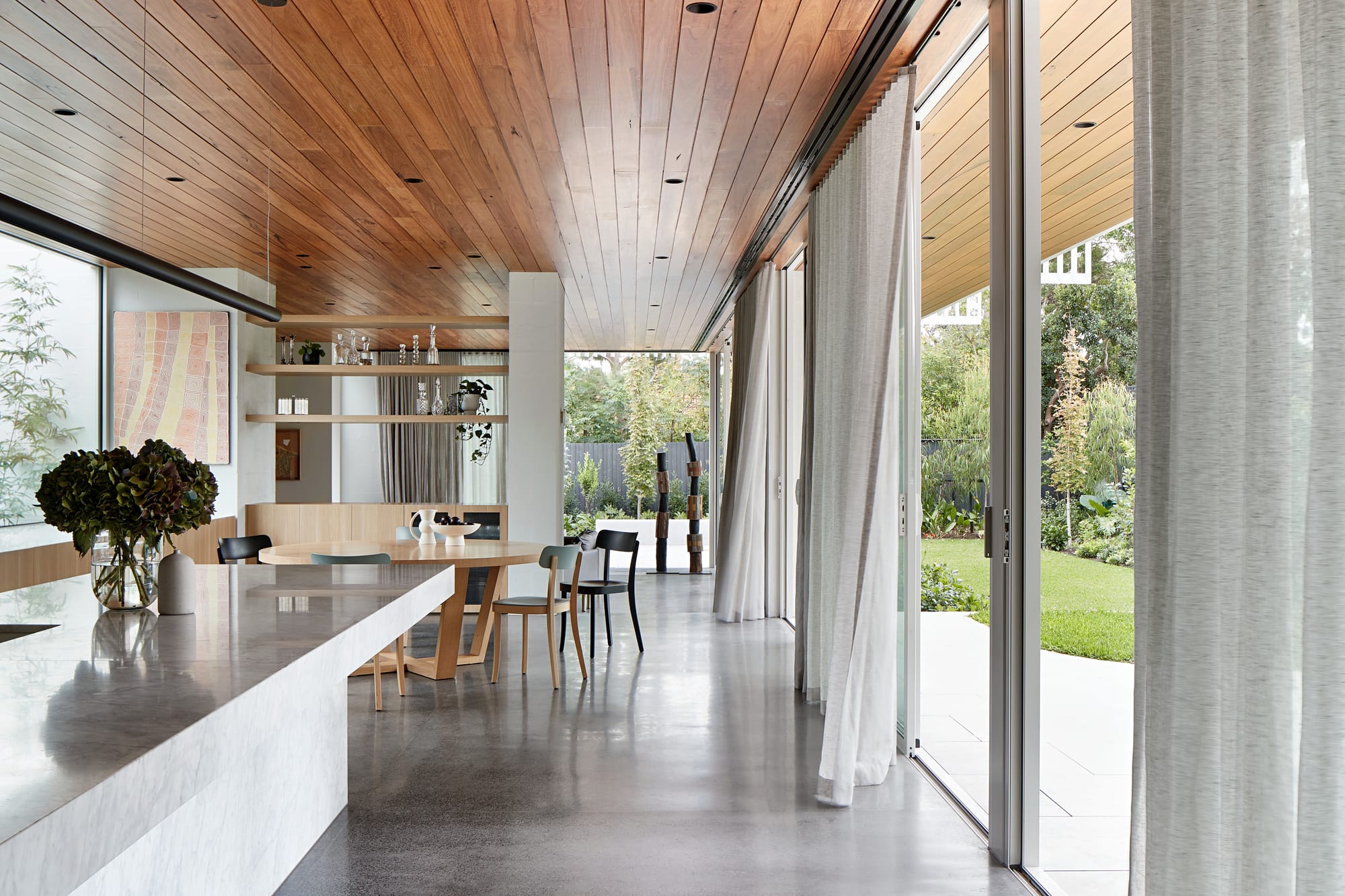 Light filled living space with a timber lined ceiling, polished concrete floors and a round timber dining table, opening onto a garden through floor to ceiling glass doors and sheer curtains.