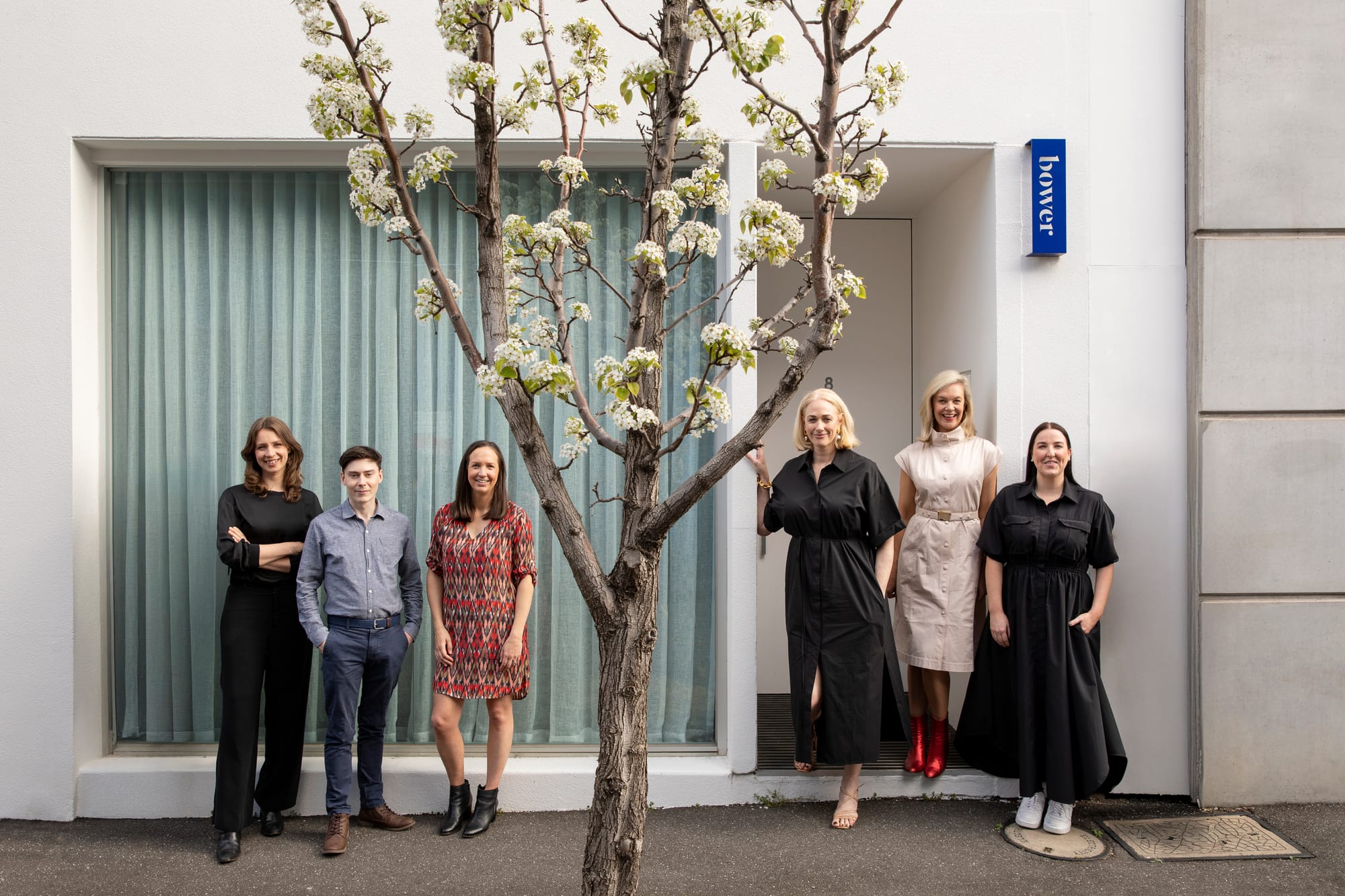 Six people standing outside a building entrance marked "bower", beneath a flowering tree, on a footpath.