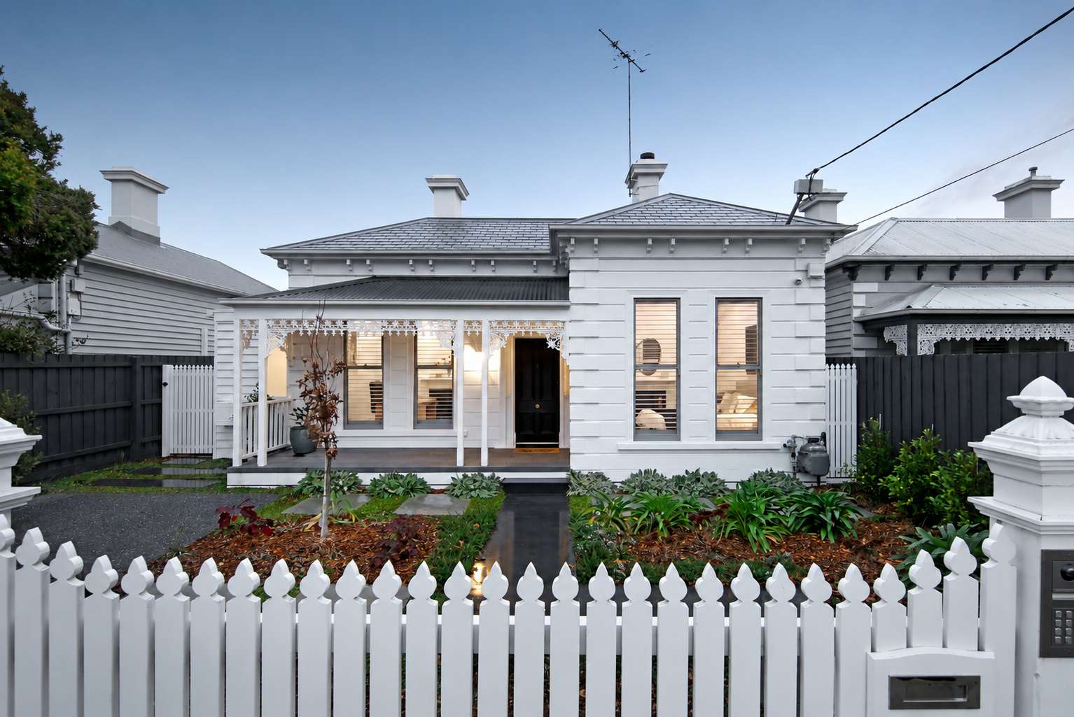 White weatherboard Victorian villa at dusk, with a lace-detailed front veranda, timber picket fence and warmly lit windows.