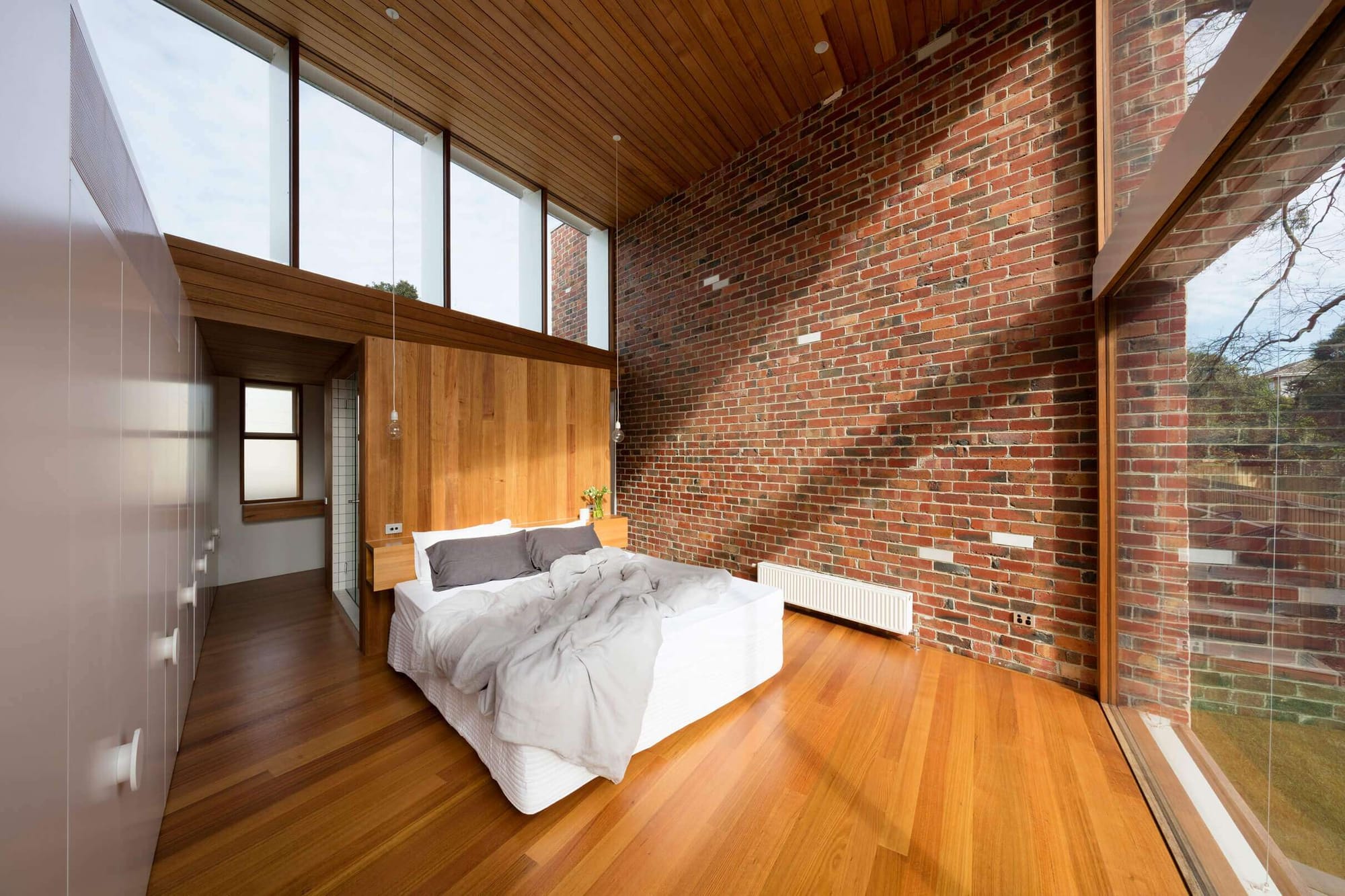 A dramatic bedroom with an exposed red brick feature wall, warm timber ceiling and wall panelling, a low platform bed with white linen, clerestory windows, and full-height glazing to the garden.