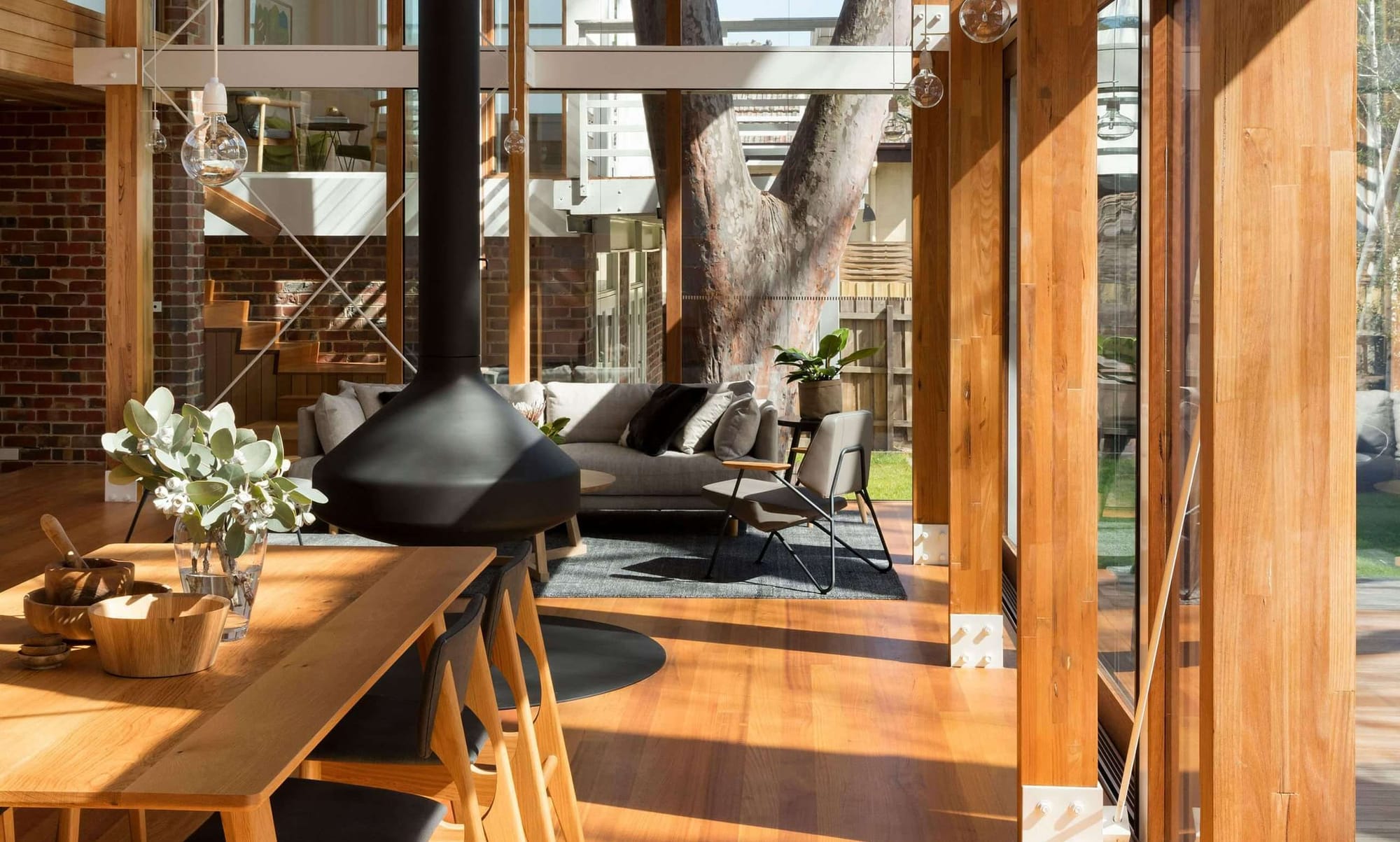 A warm timber and glass interior with a suspended black cone fireplace, timber dining table, and a view through to a living area and garden beyond, with a large mature tree visible through the glazed walls.