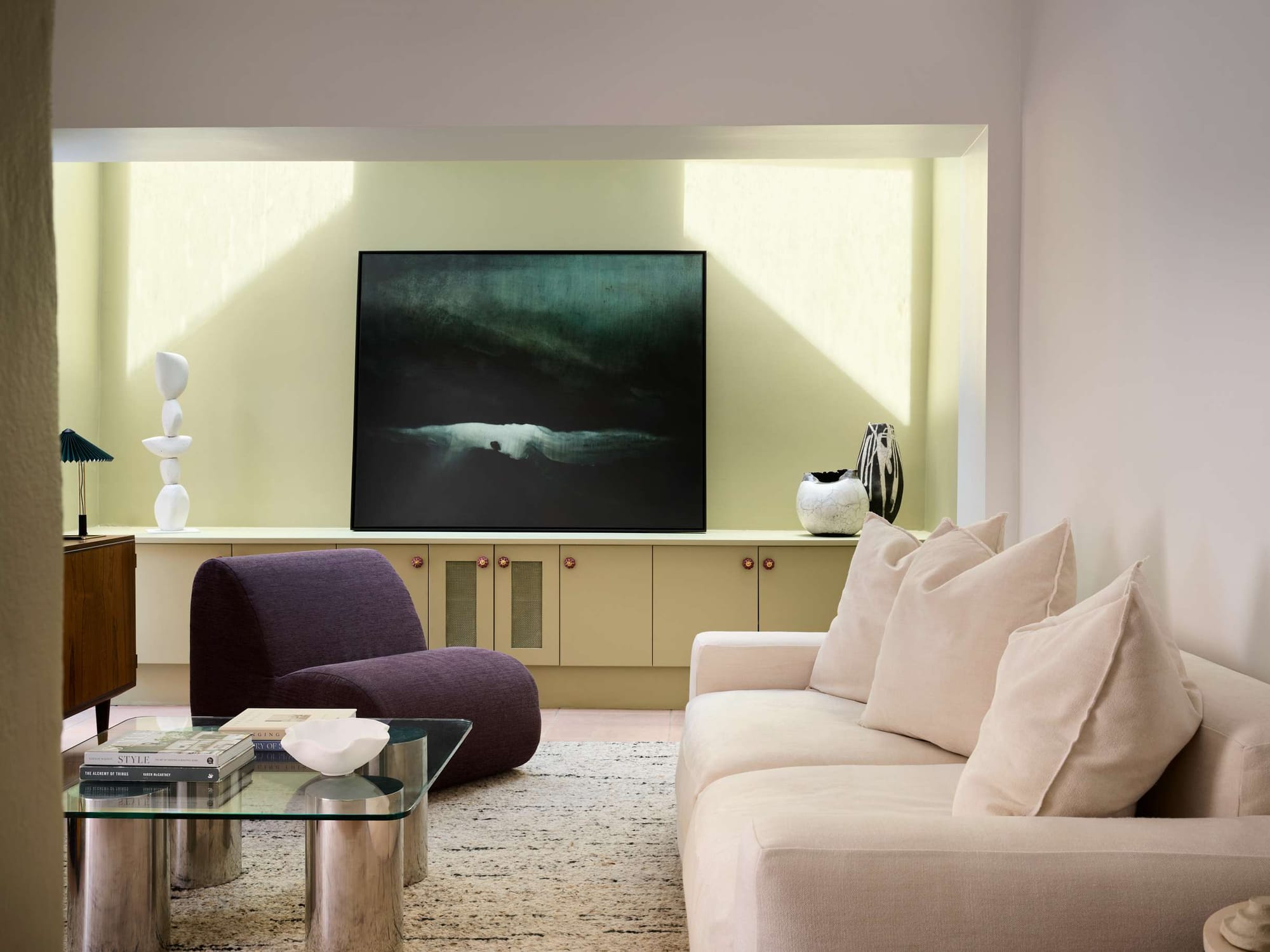 A living room with a pale yellow built-in media cabinet housing a large dark seascape artwork, flanked by white sculptural ceramics and vessels, with a curved plum accent chair, cream sofa, and glass and chrome coffee table on a textured natural rug.