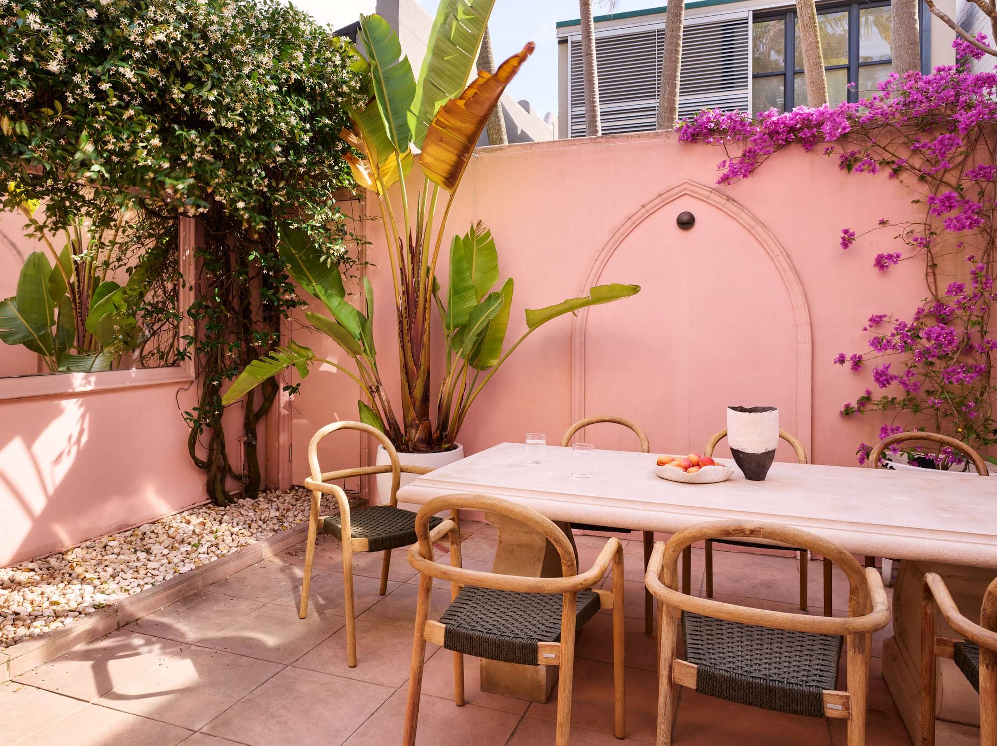 A sun-drenched courtyard with pink rendered walls, a stone dining table surrounded by timber and woven outdoor chairs, lush banana palms, climbing jasmine, and cascading pink bougainvillea.