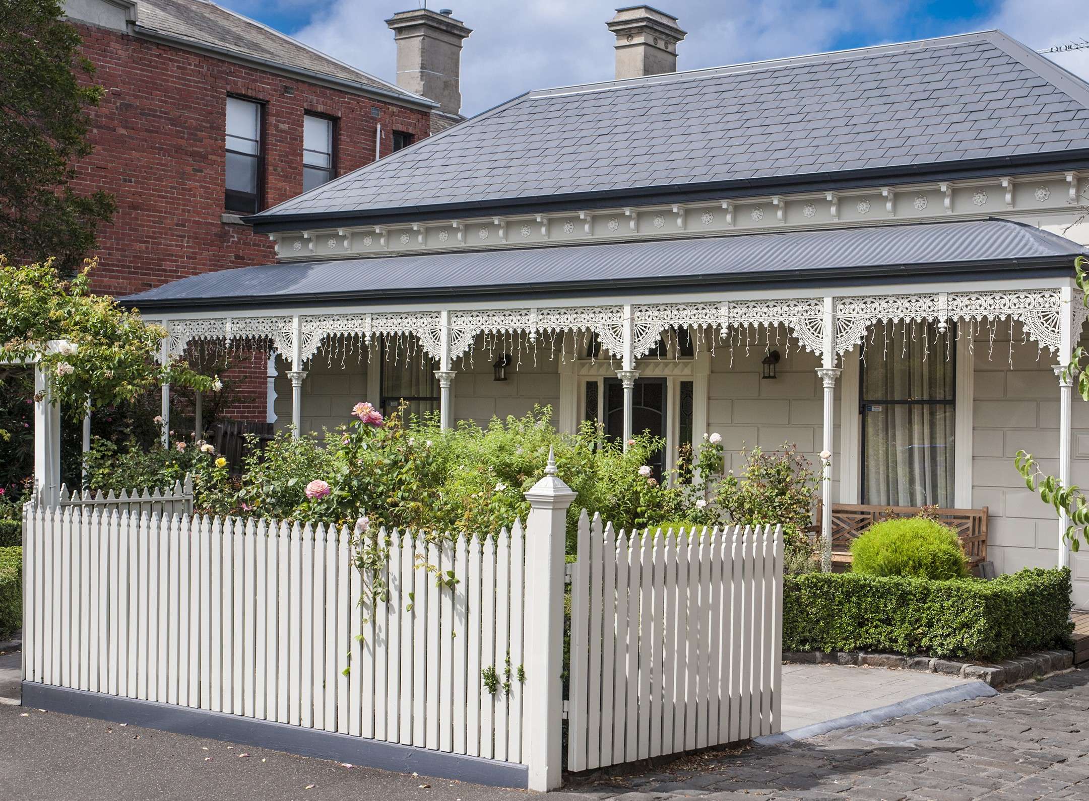 Victorian terrace home with an ornate iron lace veranda, twin chimneys and a rose-filled front garden behind a white picket fence.