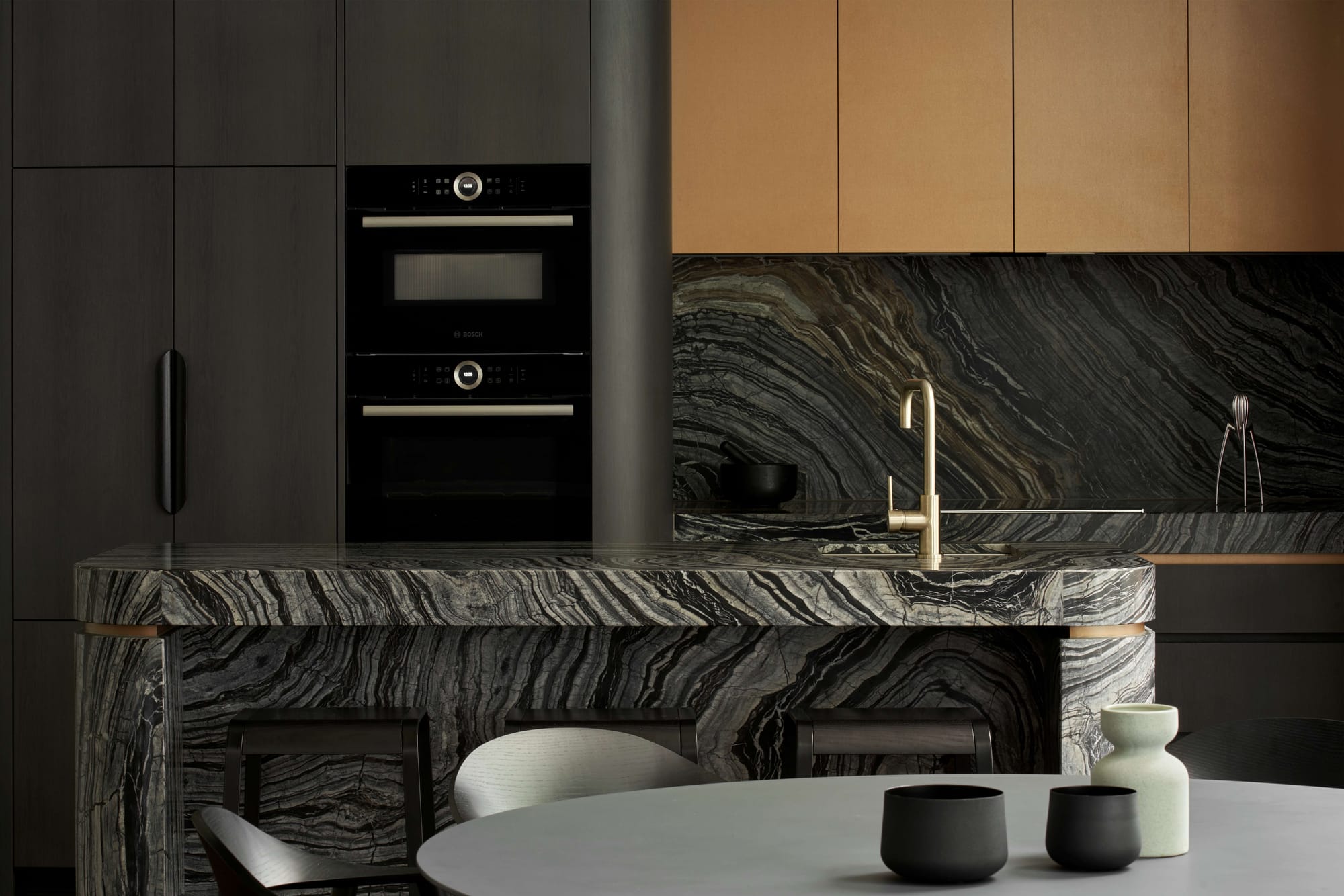 Dark marble kitchen island with a brass tap, timber upper cabinetry, and a round dining table with ceramic vessels.