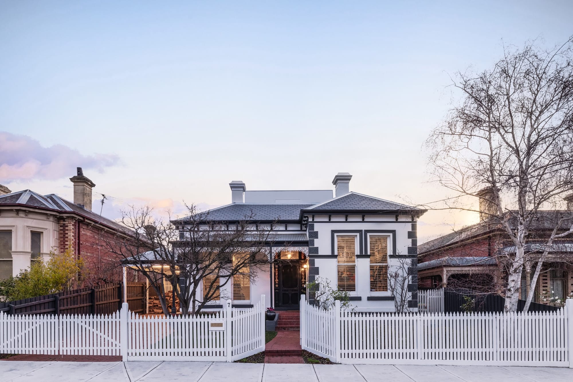 Double-fronted Victorian home at twilight with a hidden contemporary addition visible behind the original roofline, framed by a white picket fence.