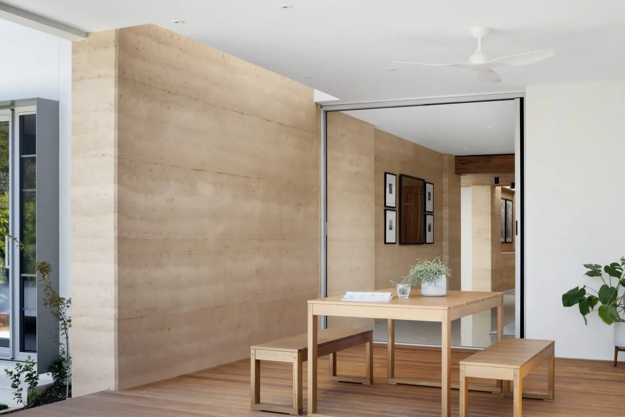 Outdoor dining area beside a rammed earth wall, a simple timber table and benches on a timber deck, with a mirrored sliding door reflecting a hallway of framed artwork inside.