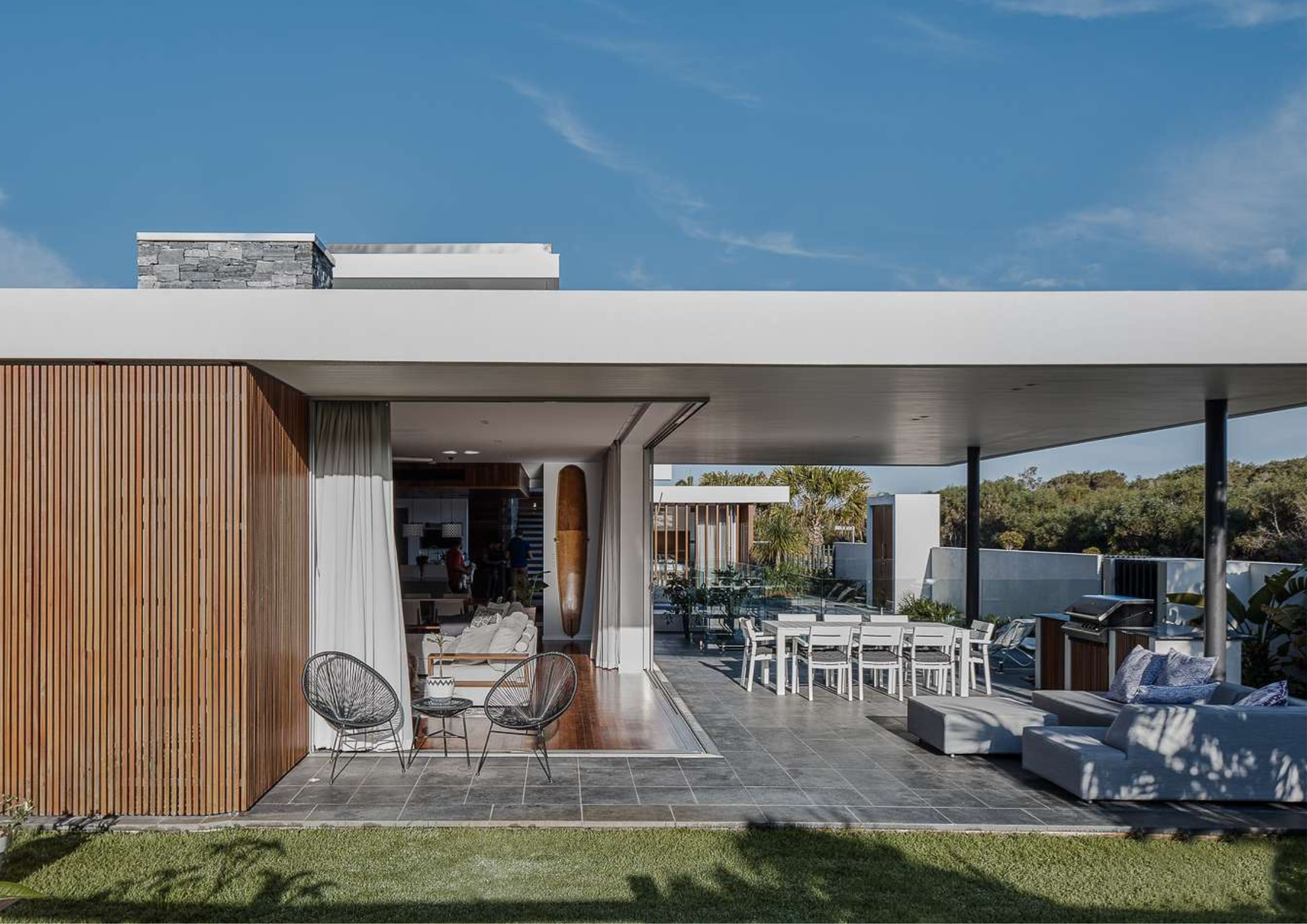 Timber-clad beach house with sliding glass doors opening to a covered alfresco with dining table, lounge seating, and built-in barbecue, overlooking a lawn and garden.