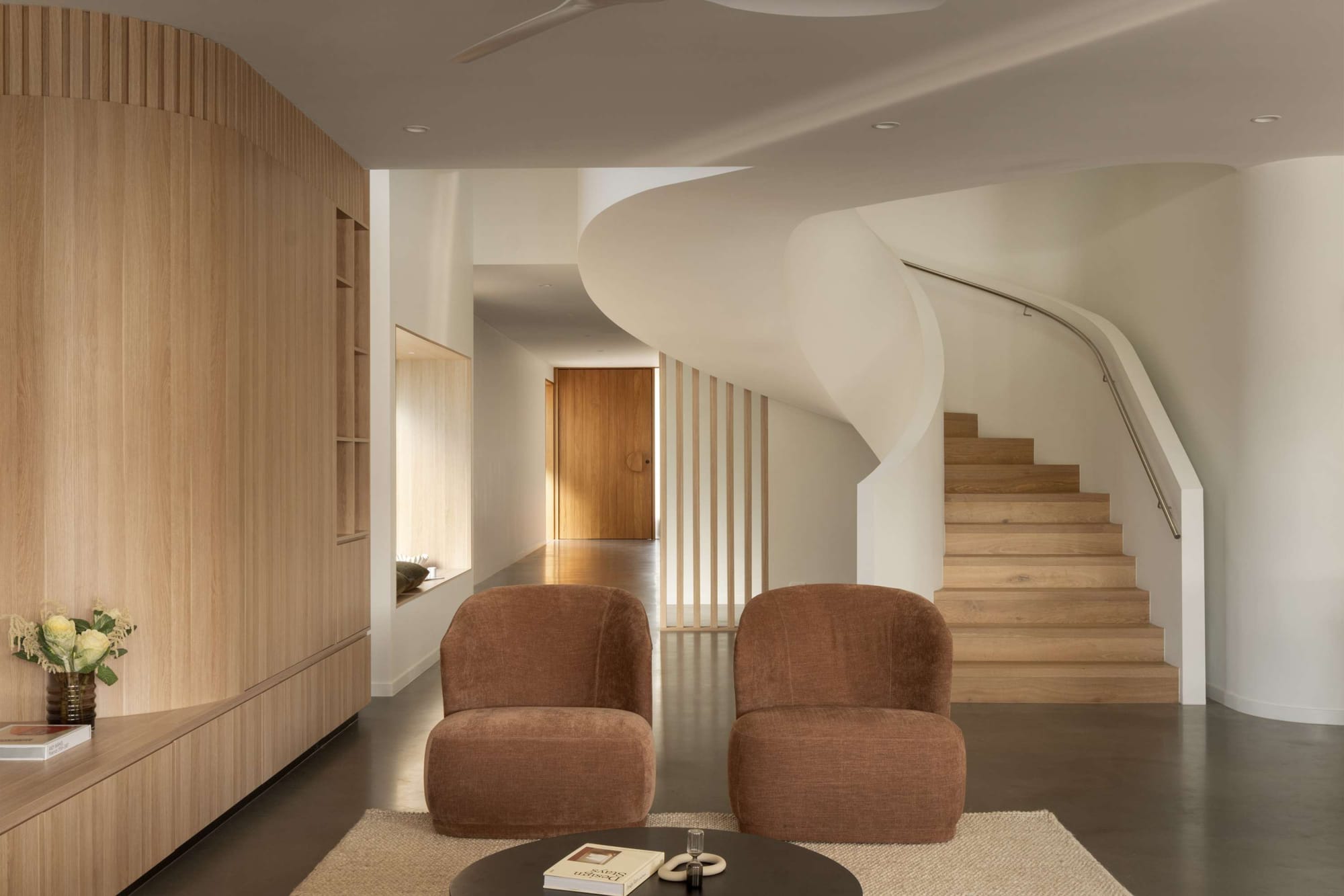 Entry hallway with a sculptural curved staircase in white plaster and timber treads, oak panelled joinery, two rust velvet armchairs and a round black coffee table in the foreground.