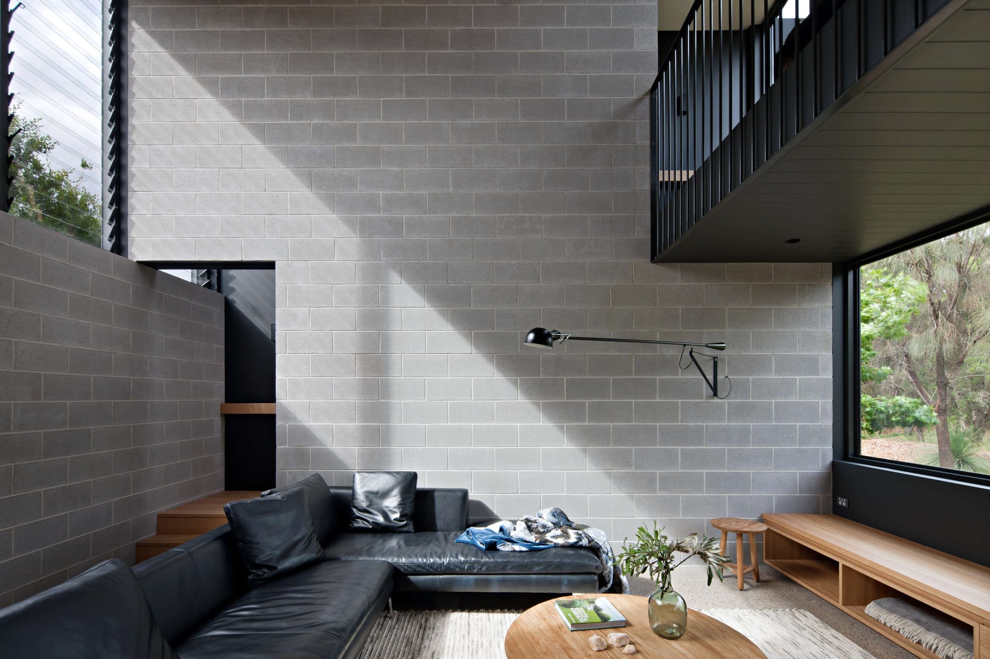 Living room with a double height grey brick wall, a black leather corner sofa and a round timber coffee table, a black articulated wall lamp and a window framing surrounding trees.