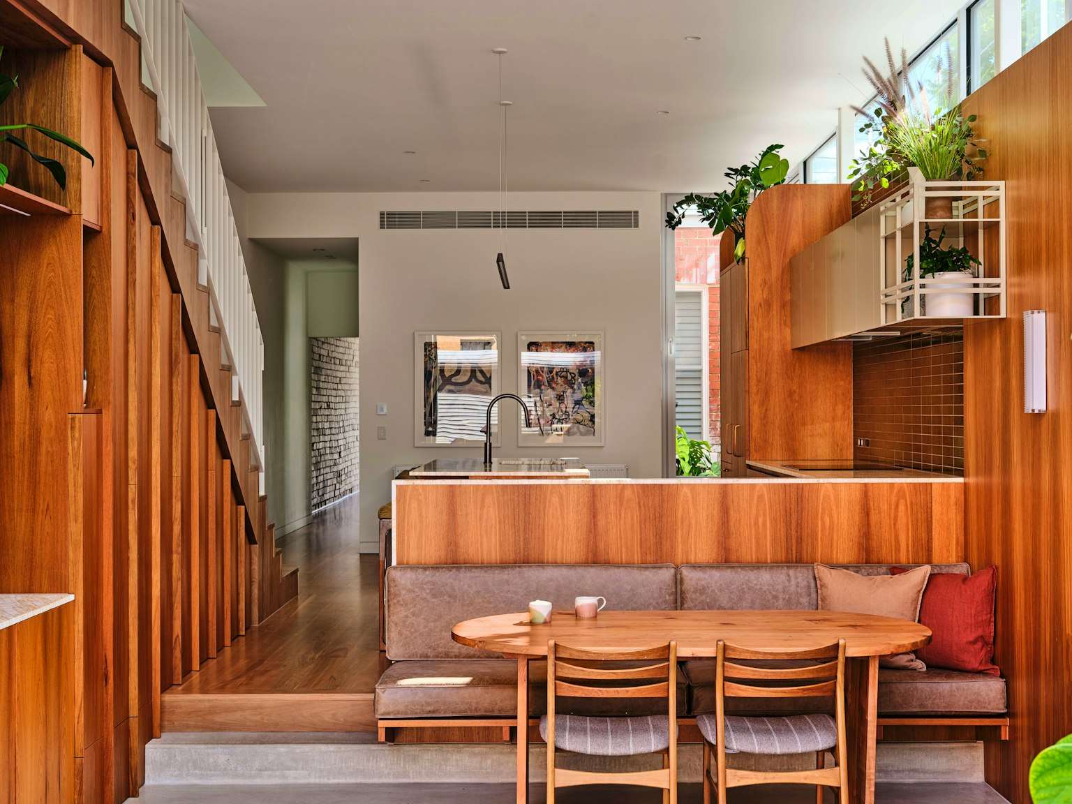 A warm timber-lined kitchen and dining area with a built-in banquette, oval timber table, island bench with stone top, staircase with integrated shelving, and plants cascading from upper shelves.