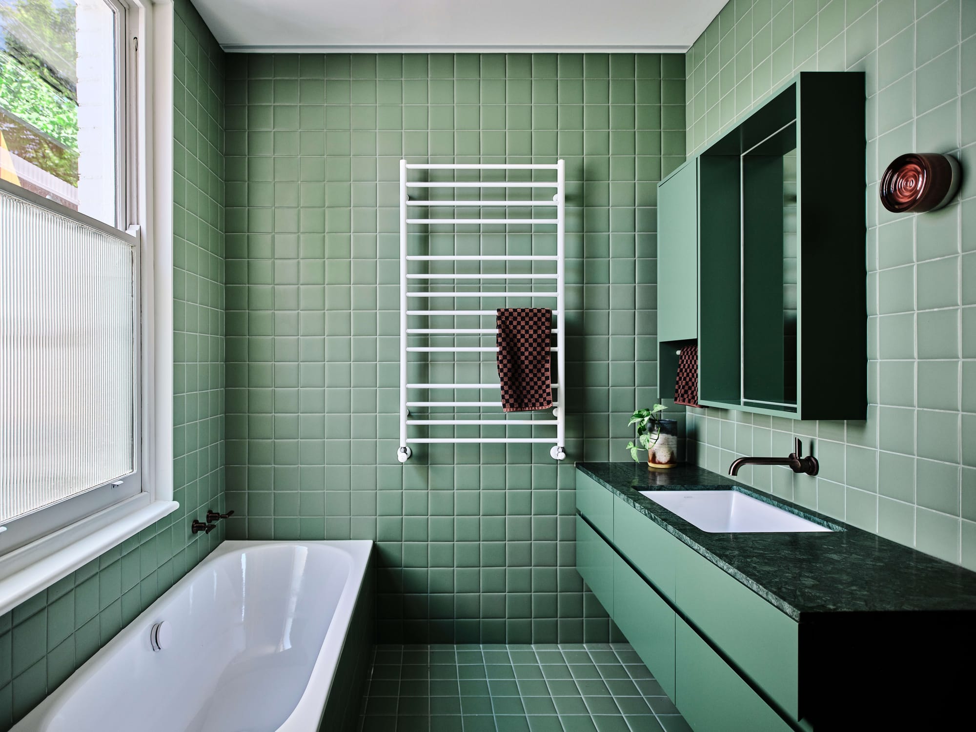 A fully tiled bathroom in sage green with a freestanding bath, white ladder towel rail, dark green marble vanity, recessed mirror cabinet, and a ribbed glass window.