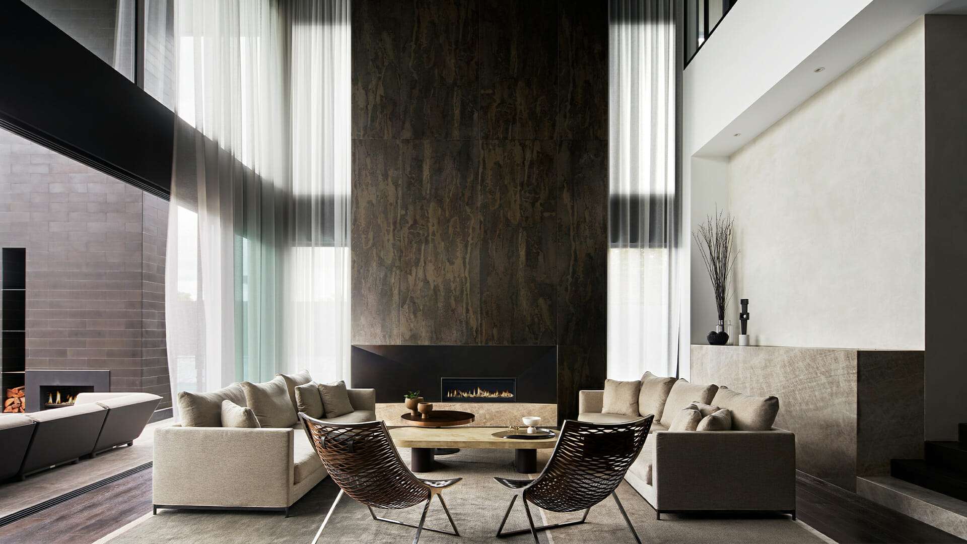Double height living room with a textured bronze fireplace wall, sheer curtains, and woven leather accent chairs.