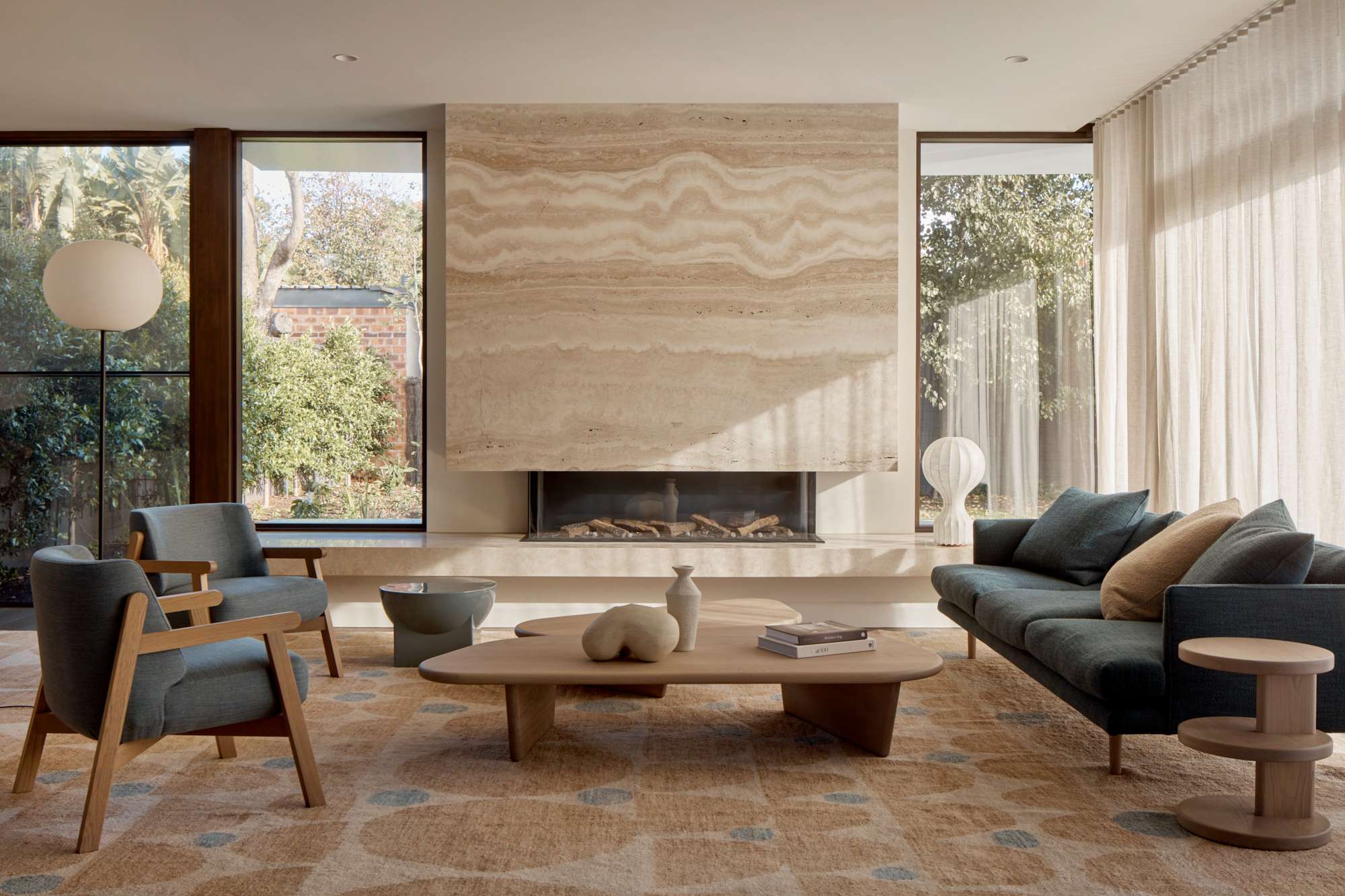 Living room with a floor to ceiling travertine fireplace surround, timber coffee tables, and blue upholstered armchairs on a patterned rug.