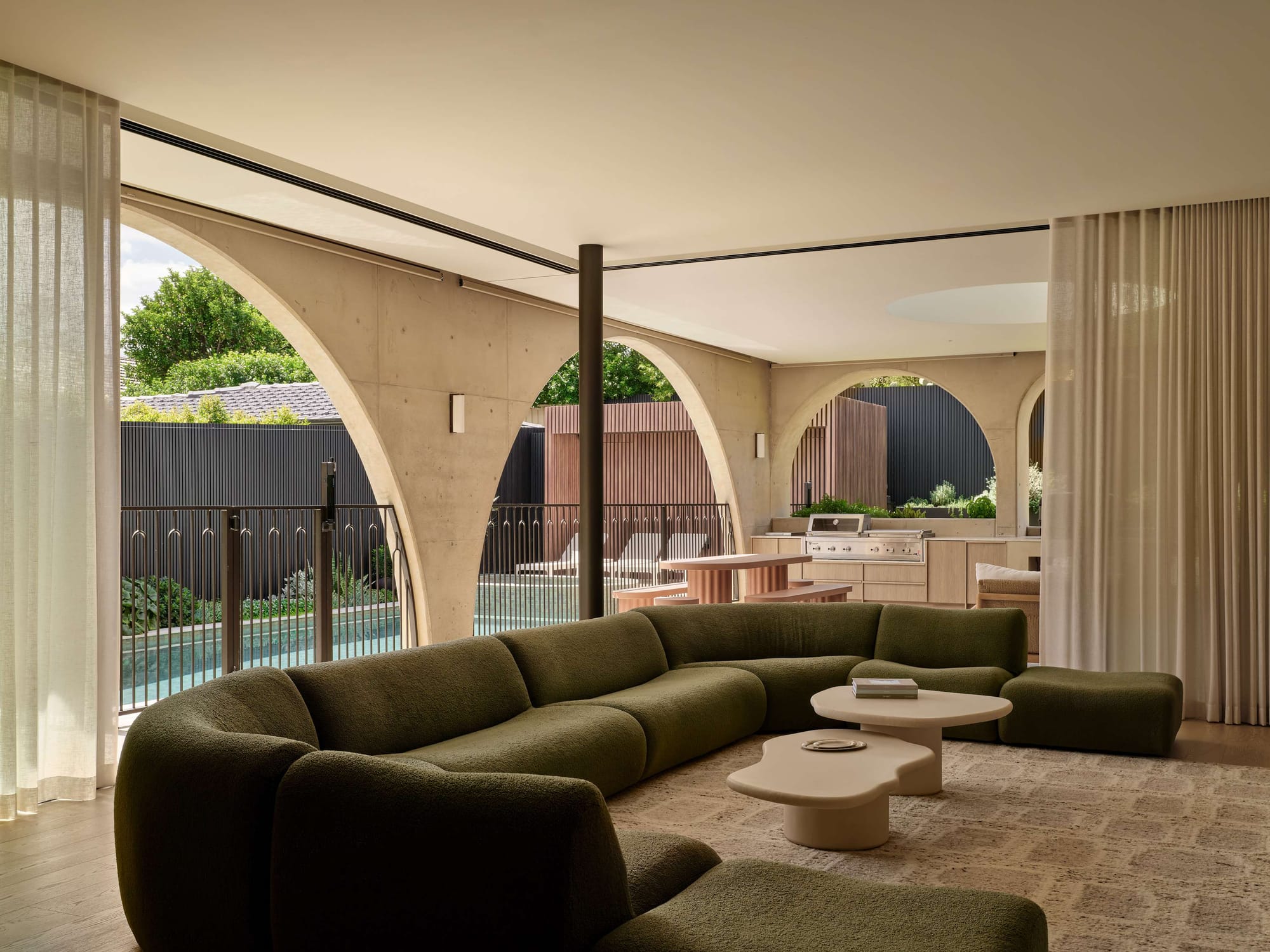 Outdoor living pavilion with concrete arches, sheer curtains, and an olive green curved sofa overlooking a pool.