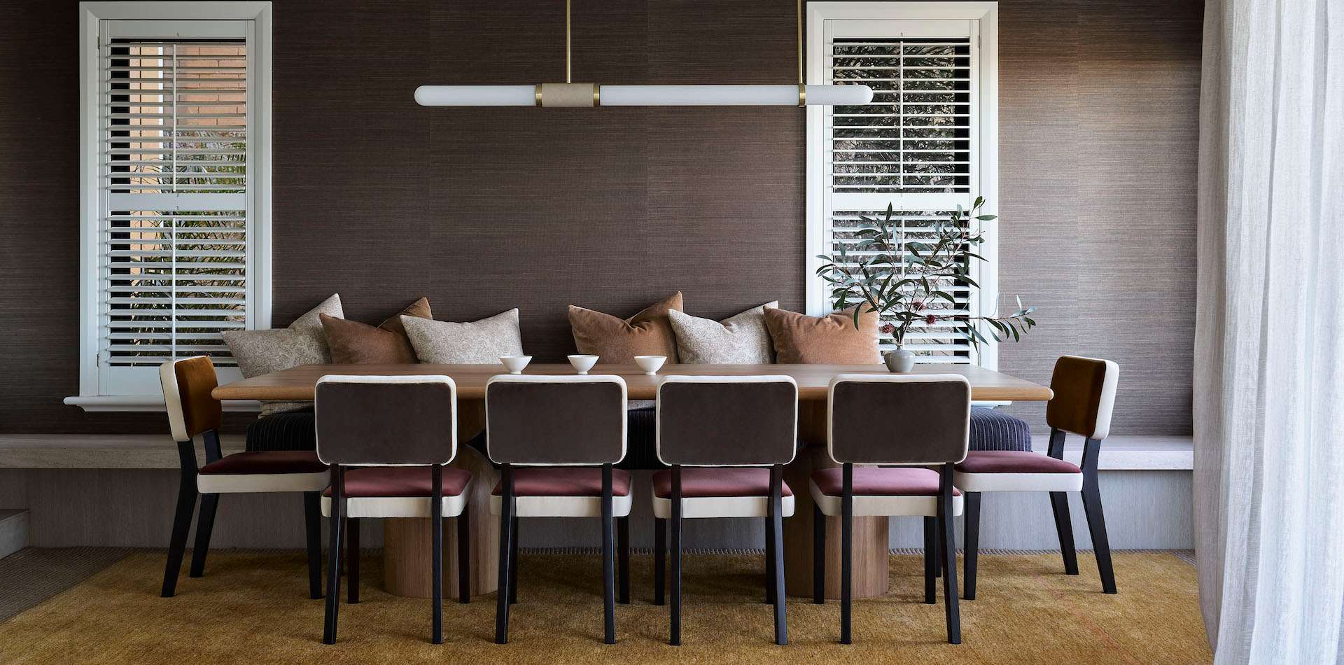 Dining room with a built in banquette seat and dark grey textured wallpaper, timber dining table surrounded by upholstered chairs in burgundy and cream, brass pendant light suspended above. Interior design by Marylou Sobel Interior Design. Photography by Anson Smart.