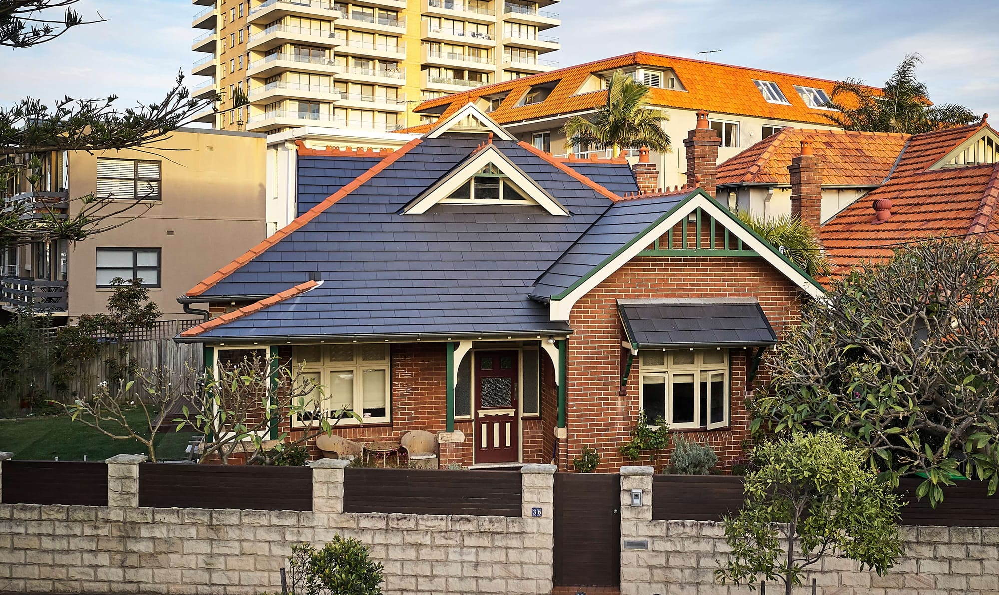 Federation-era red brick bungalow with a new slate roof and timber gable detailing, set against a backdrop of surrounding apartment towers.