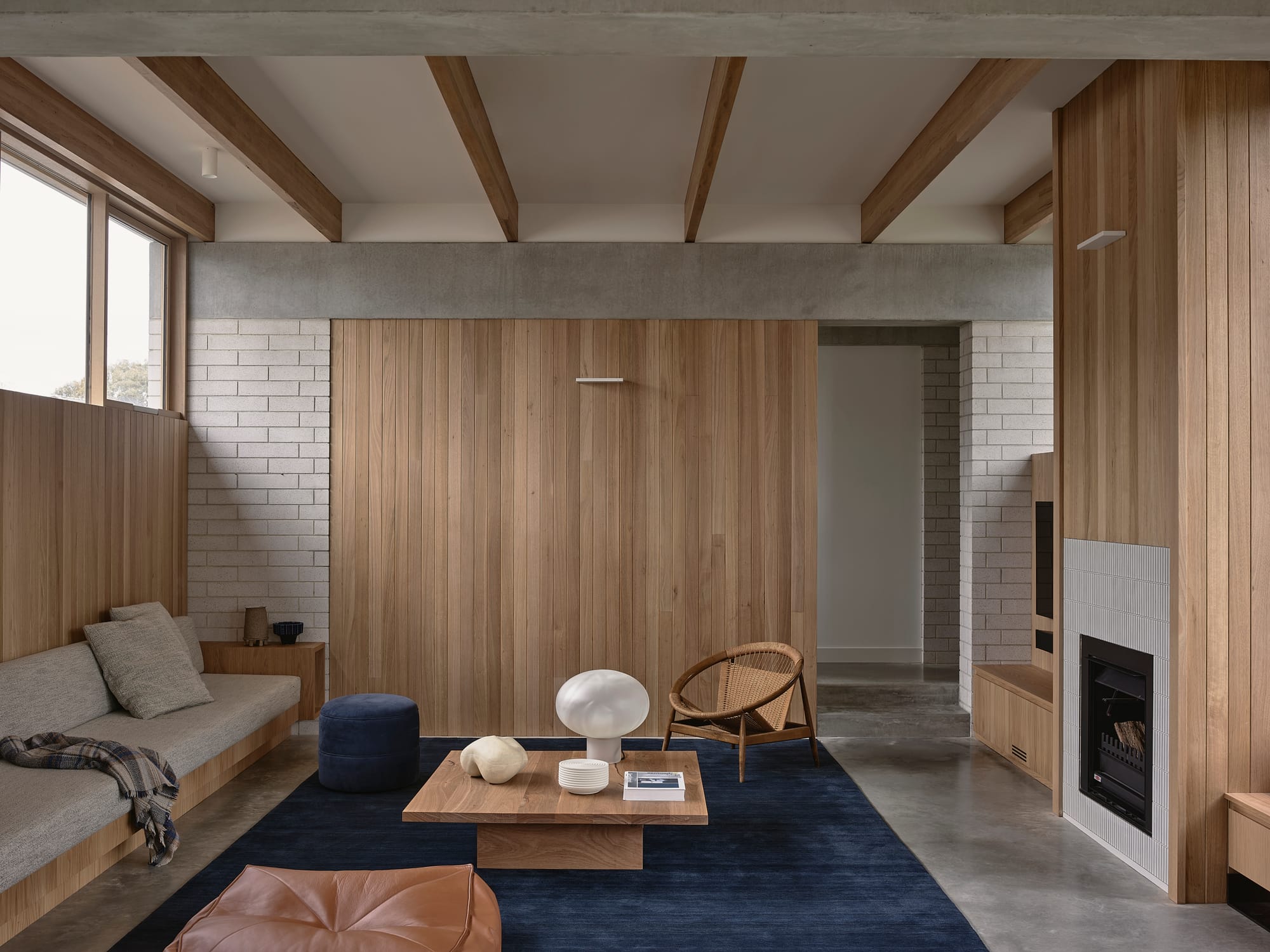 A warm living room interior with vertical timber wall panelling, exposed concrete beams, grey brick walls, polished concrete floors, a built-in timber sofa, low coffee table, and a gas fireplace set into a ribbed tile surround.