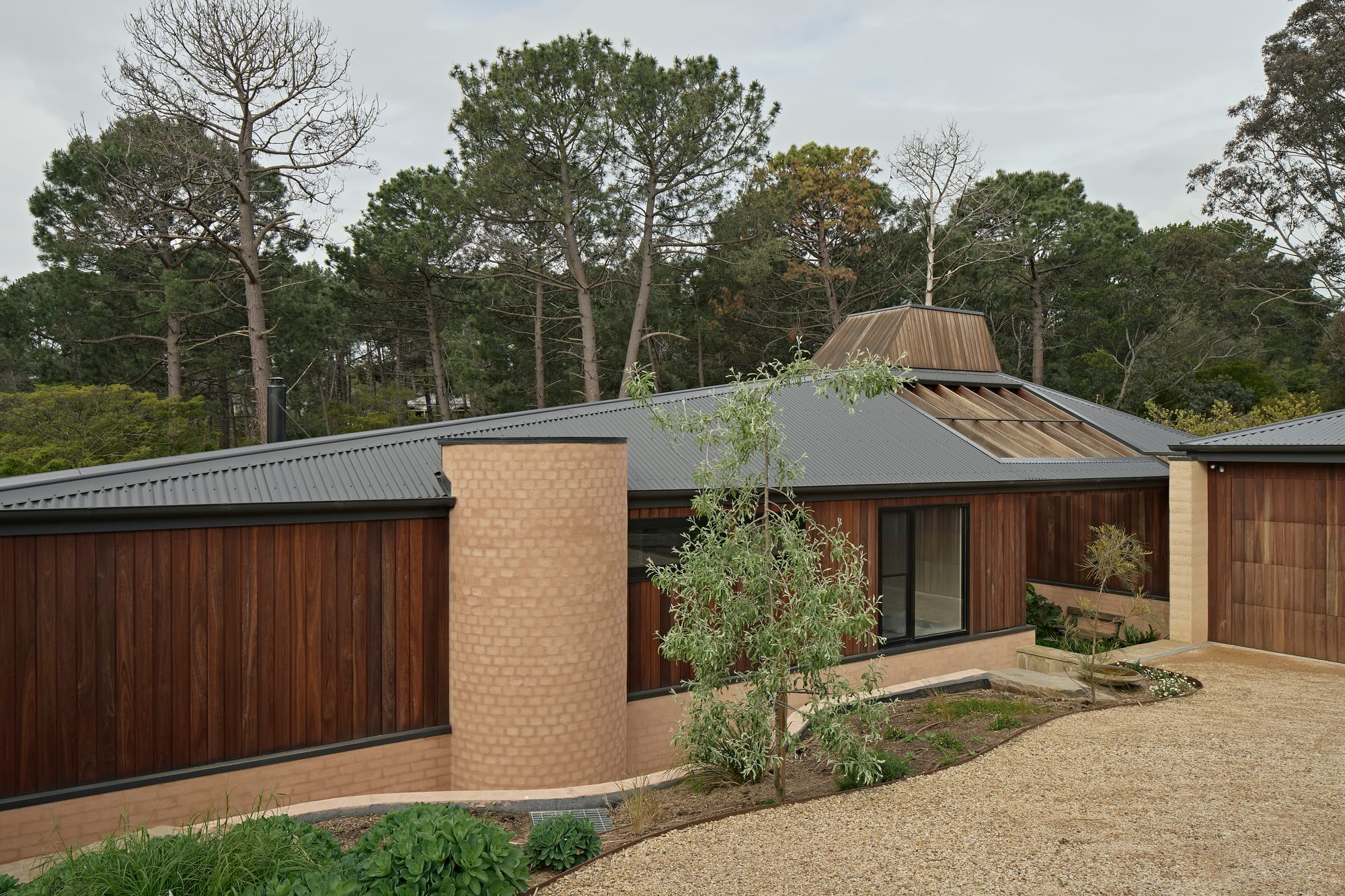 The exterior of a low-set home with warm brick masonry piers, dark timber cladding, a corrugated iron roof, and a distinctive copper-clad lantern skylight rising from the roofline, set against a backdrop of tall pine trees.