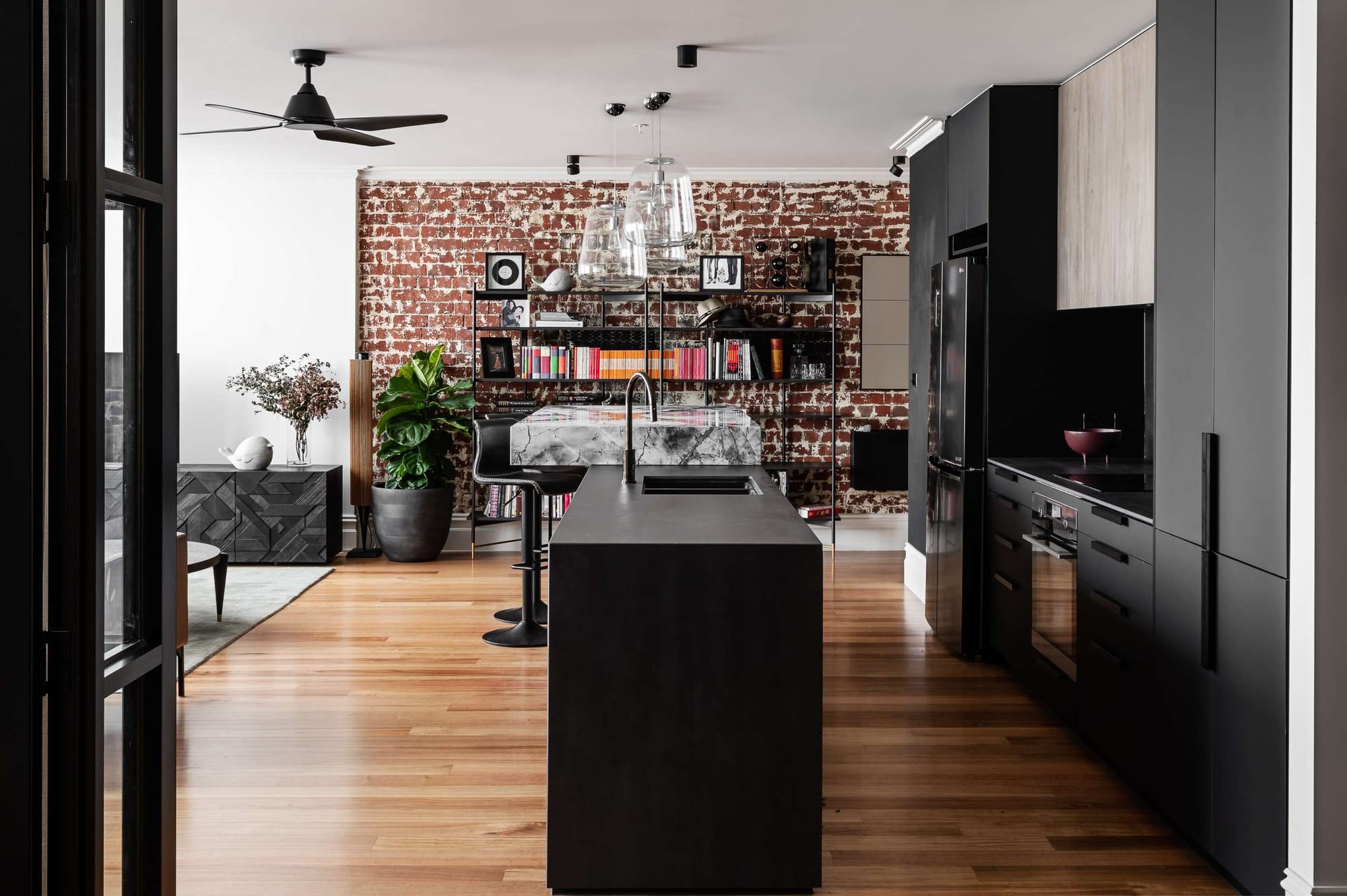 Open plan kitchen with a black stone island, timber flooring, and a distressed brick wall lined with open shelving.